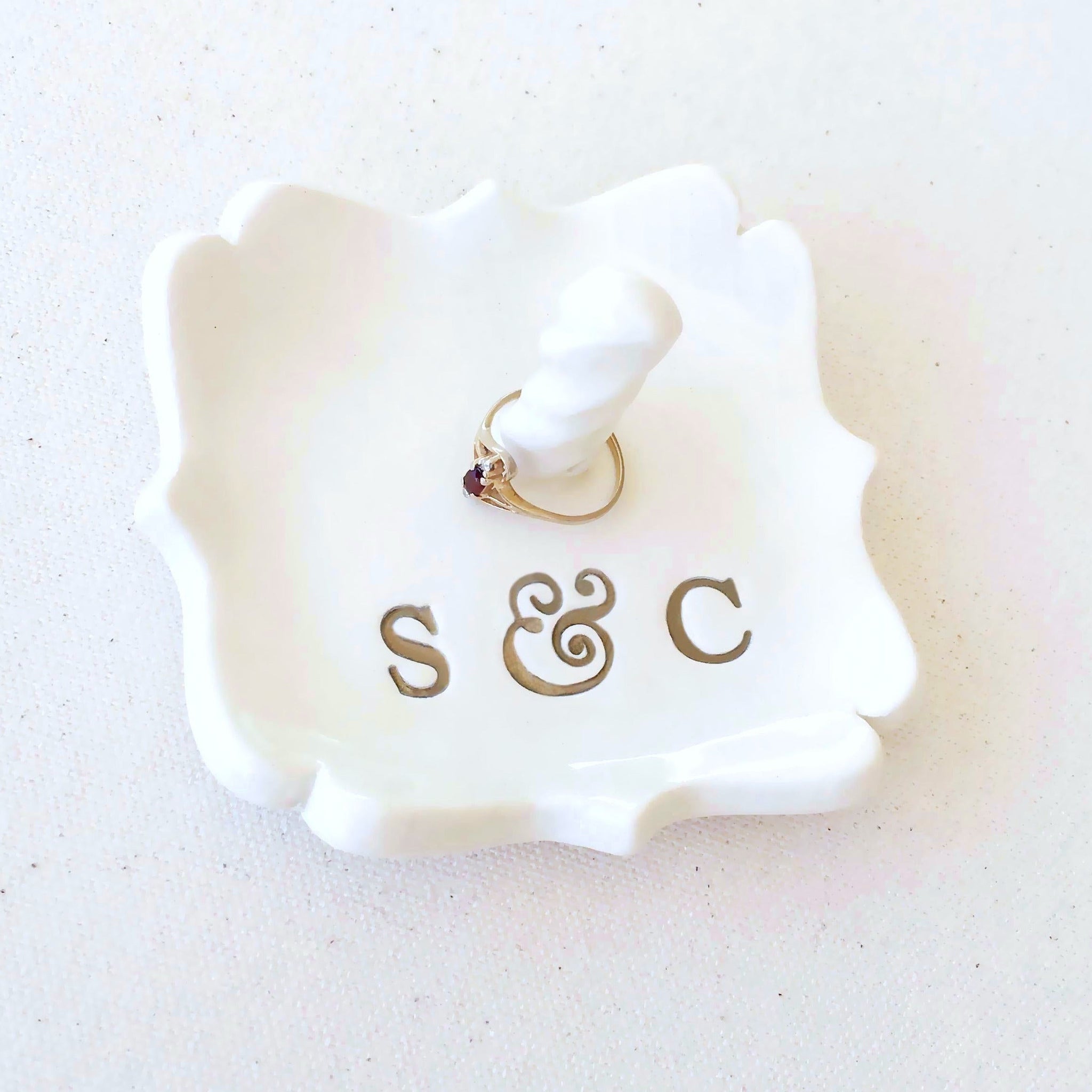 A white ceramic ring holder with scalloped edges, gold monogram letters 'S & C', and a finger ring displayed on its jewelry post, dish is set against a light textured background. Handmade ceramic personalized ring cone dish, custom pottery white square tray with scalloped curvy sides has custom typographic design includes personalization with 2 name initials. Ideal wedding ring tray. Initials are painted gold. Perfect custom wedding anniversary gift spouse anniversary present.