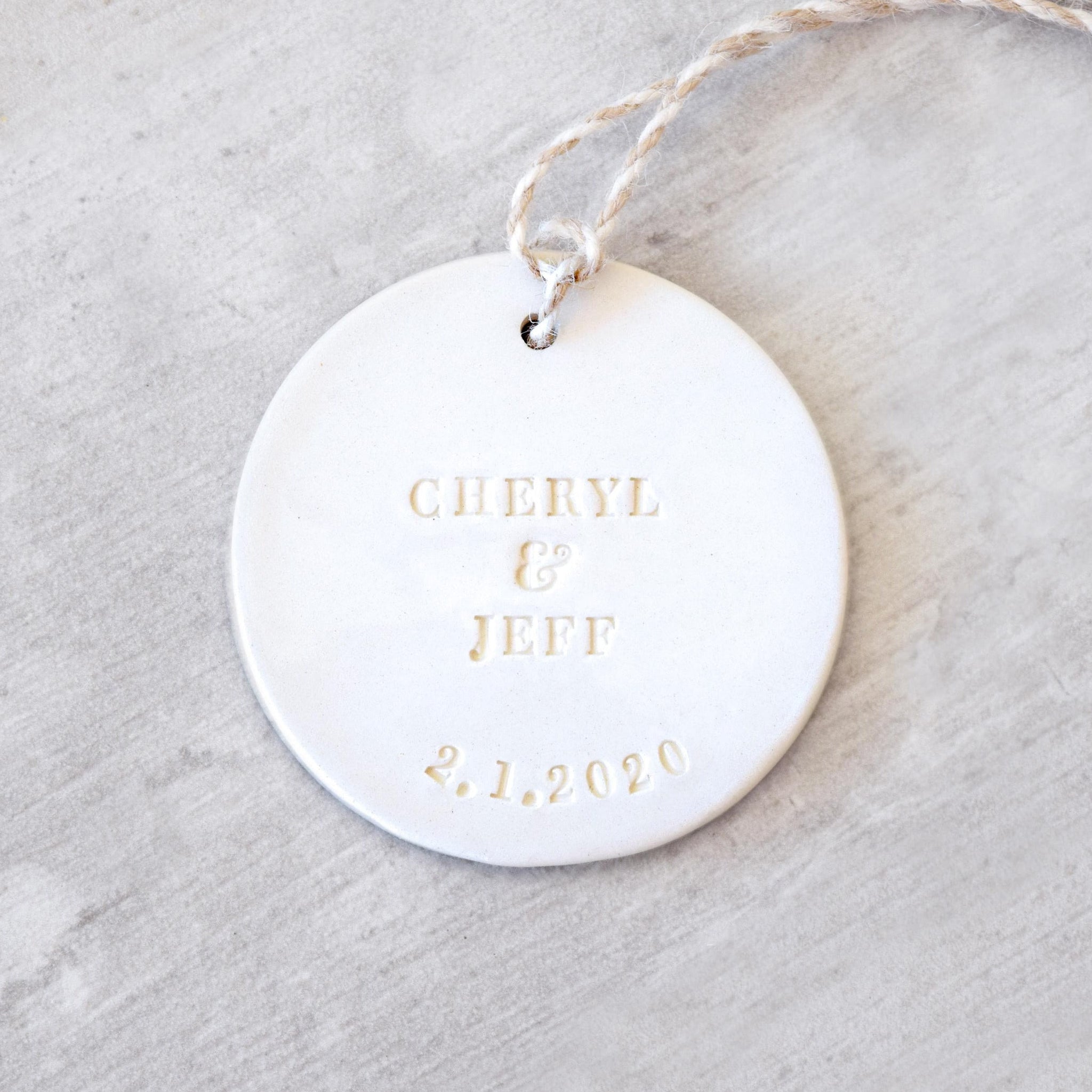 Handmade ornament ceramic clay charm white round flat disc with smooth surface, tan hang cord and typographic design in classic letter style on gray surface.  Personalized weddings engagement keepsake created with 2 first names and a date. Custom pottery ornament hanging tree medallion, weddings keepsake for country farmhouse minimalist holiday decor marriage or couple proposal gift.