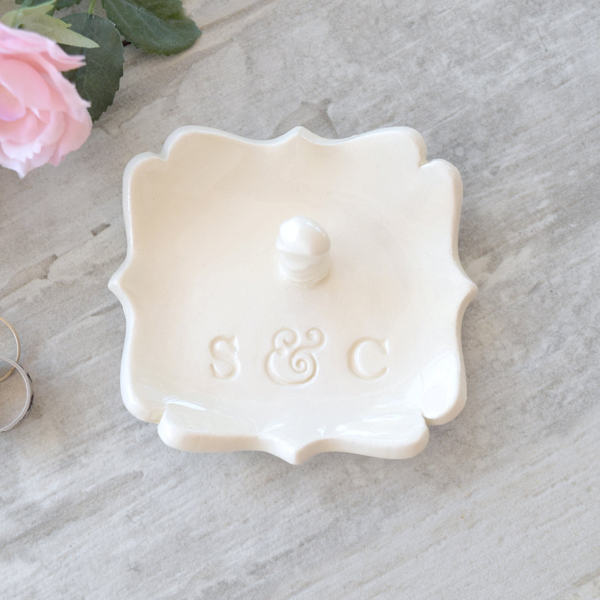 A white ceramic ring holder with scalloped edges, monogram letters 'S & C' stamped in clay and a 3d jewelry post, dish is set against a gray wood surface with pink flowers. Handmade ceramic personalized ring dish with post, custom pottery white square tray with scalloped curvy sides has custom typographic design includes personalization with 2 name initials. Ideal wedding ring tray.  Perfect custom wedding anniversary gift spouse anniversary present.