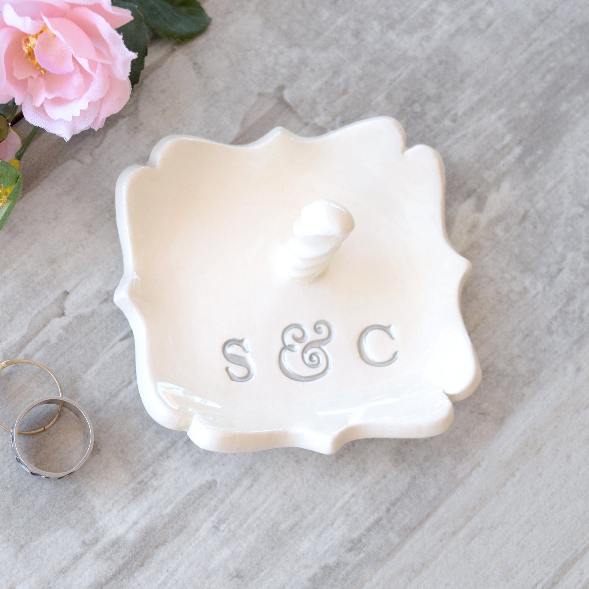 A white ceramic ring holder with scalloped edges, silver monogram letters 'S & C', and a finger ring jewelry post, dish is set against a light gray background with flowers. Handmade ceramic personalized ring cone, custom pottery white square tray with scalloped curvy sides has custom typographic design includes personalization with 2 name initials. Ideal wedding ring tray. Initials are painted silver. Perfect custom wedding anniversary gift spouse anniversary present.