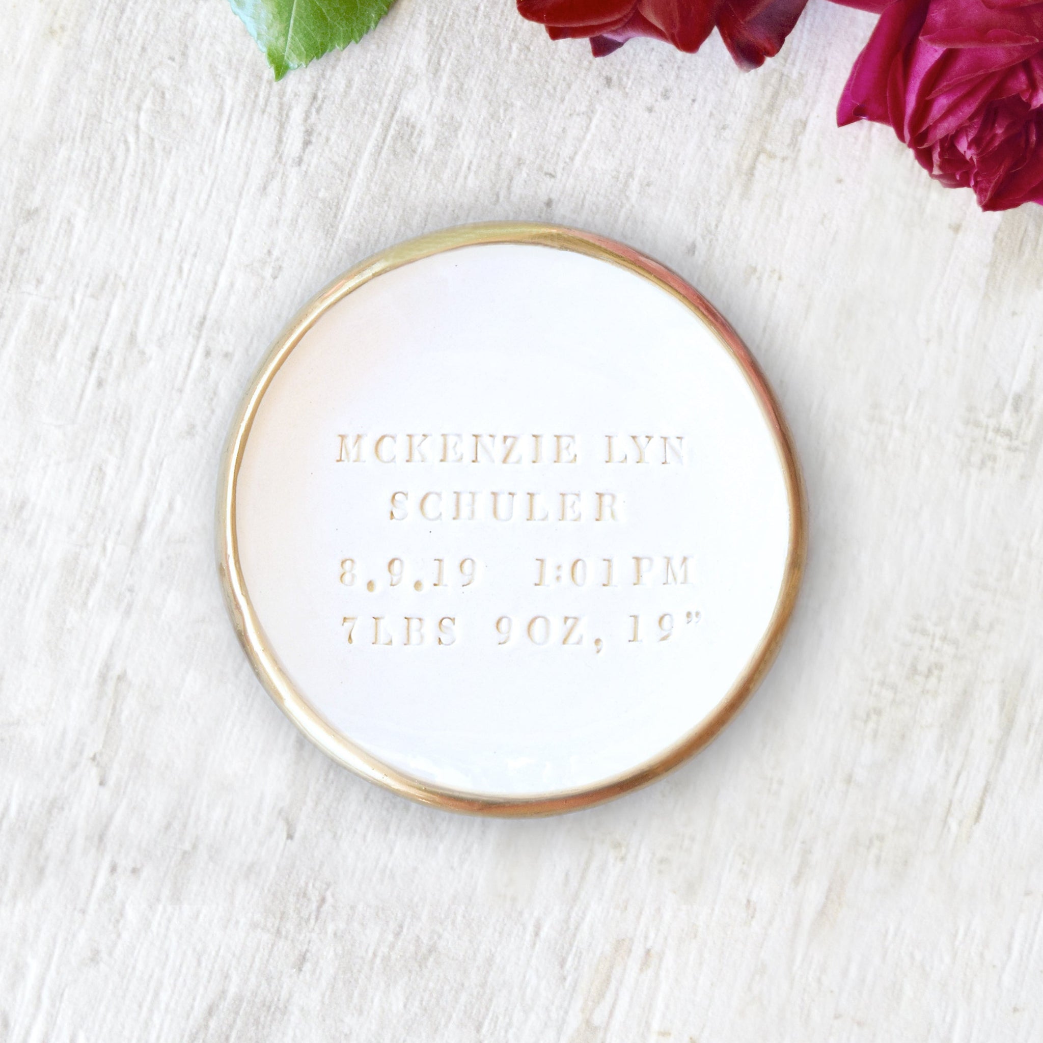 handmade ceramic gold rim white gloss bowl with typographic design hand stamped in clay on wood surface with pink flowers. Personalized baby birth specs bowl, hand engraved birthday keepsake for newborn with name date time weight length. Custom day you were born dish for new parents gift heirloom.