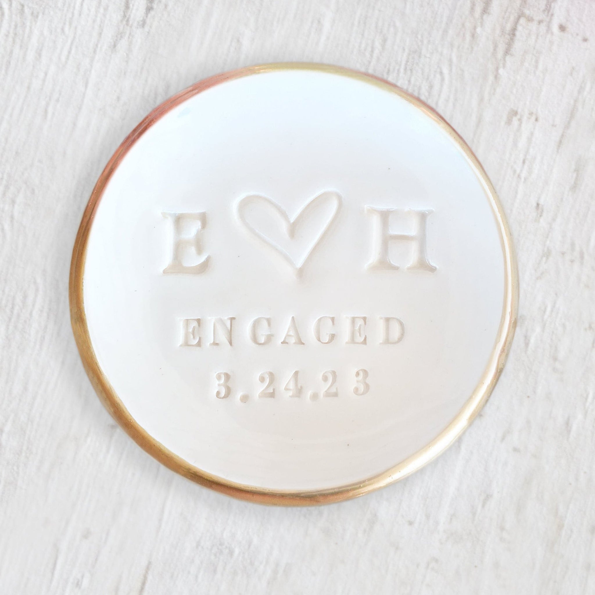 classic white bowl with gold trim on wood surface. Handmade ceramic custom engagement ring dish for engaged couple gift, design features hand engraved 2 name initials, a heart, the word engaged and a date. Future Mrs wedding jewelry holder proposal keepsake