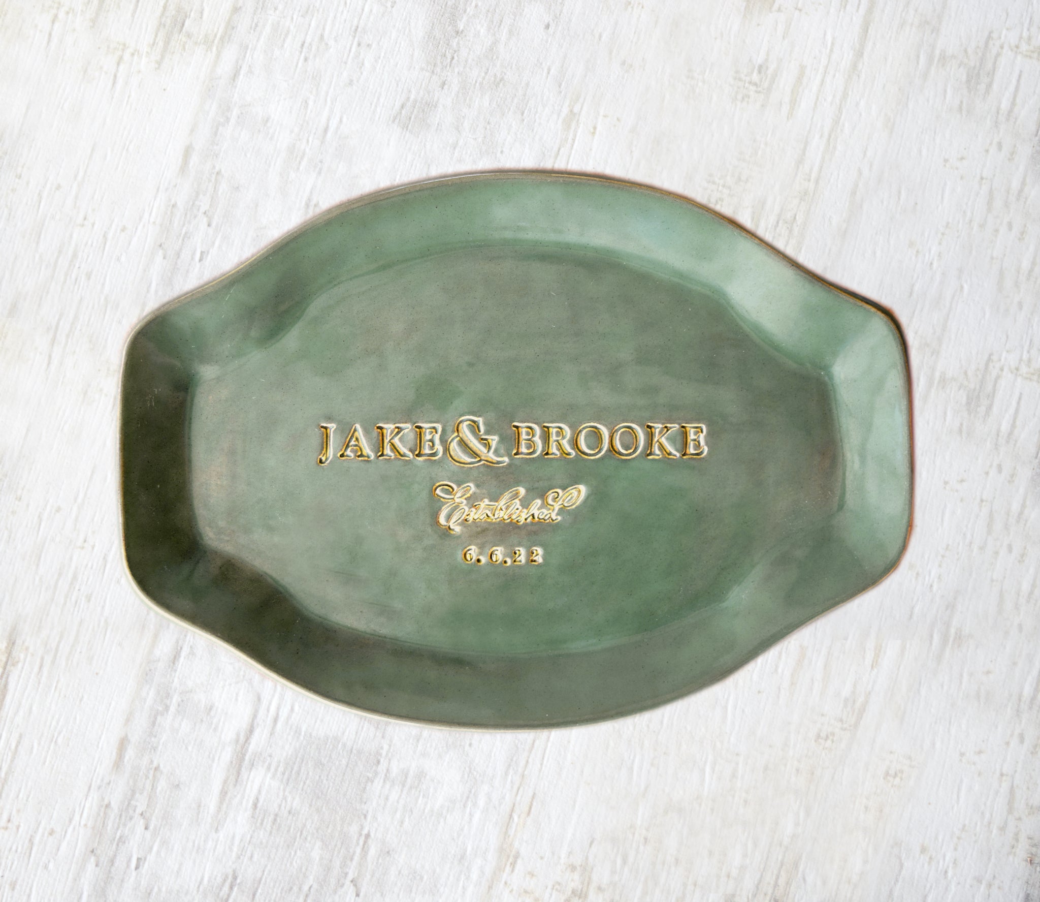 handmade ceramics rustic green oval platter. Gloss glazed personalized pottery platter with handles, hand engraved 2 names, the word established and a date, golden accents on trim and gold lettering. Dish shown on a light wooden surface. unique wedding anniversary gift for couple, ceremony plate keepsake