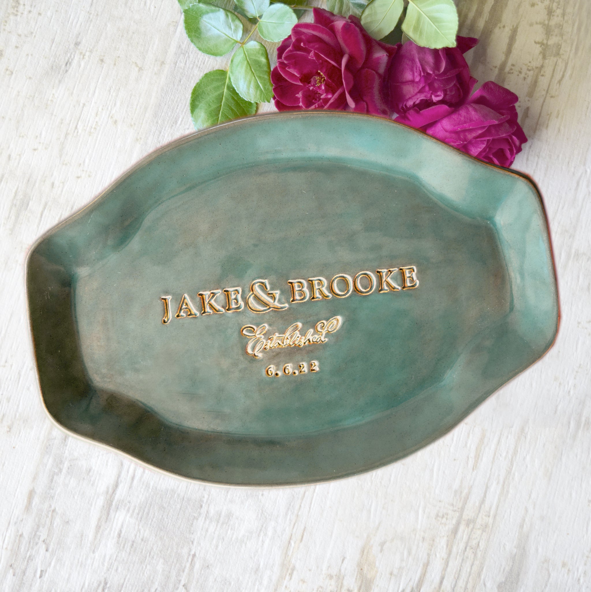 Green handmade ceramic platter with typographic design pressed in clay on wood surface with red flowers. Round serving tray Includes 2 first names, the word established and a date outlined in antique gold. Unique personalized kitchen wedding gift anniversary plate keepsake for couple, friends and family life event memorial celebration. 