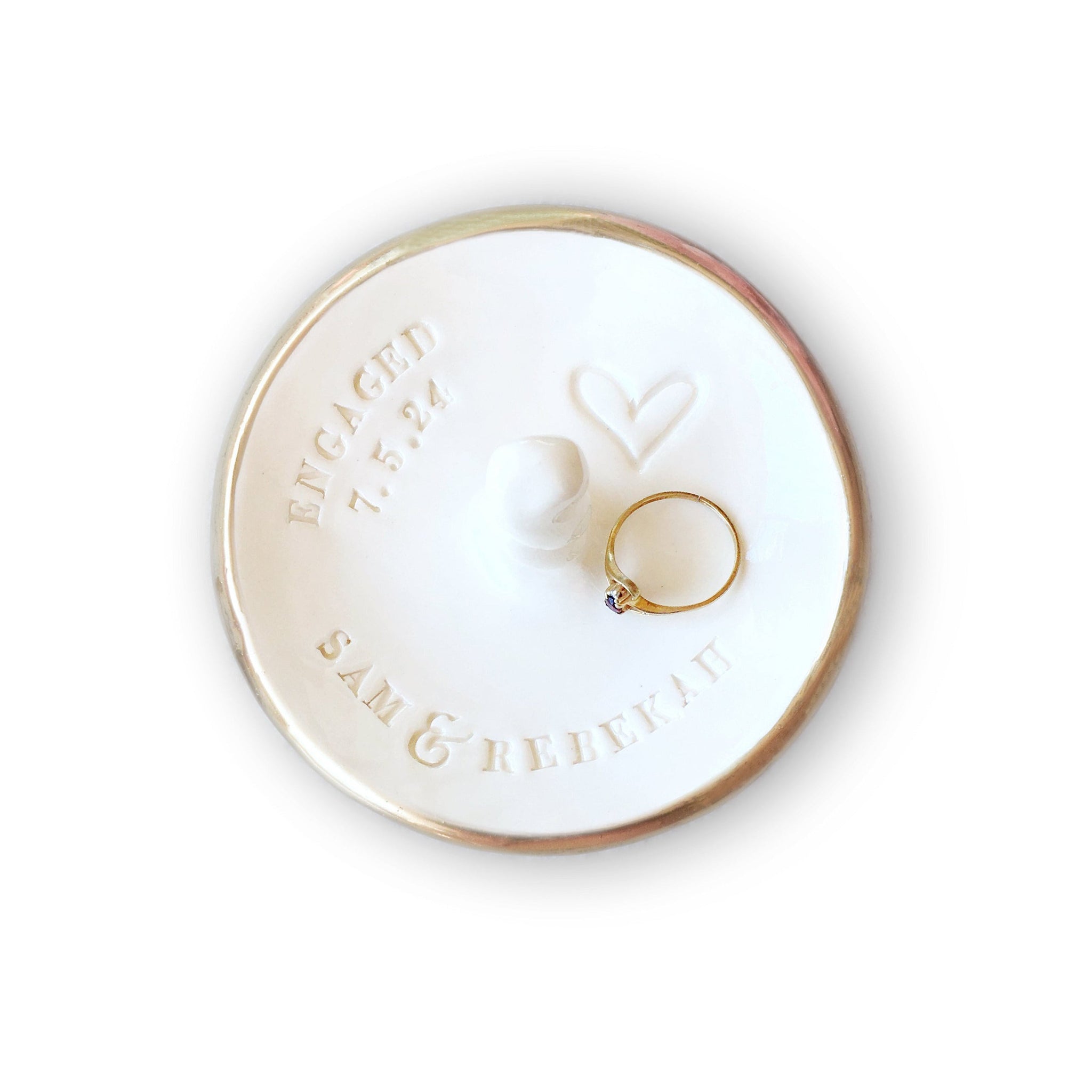 Handmade ceramic round white dish with centered rings post & gold rim edge holding a ring on a white background. Custom engagement gift personalized ring dish with post, includes hand engraved heart, 2 names & optional word engaged and date, also features a central jewelry display cone to safely secure bracelets, rings, necklaces. Newly engaged couple gift, bridal shower present, future mr and mrs wedding keepsake.