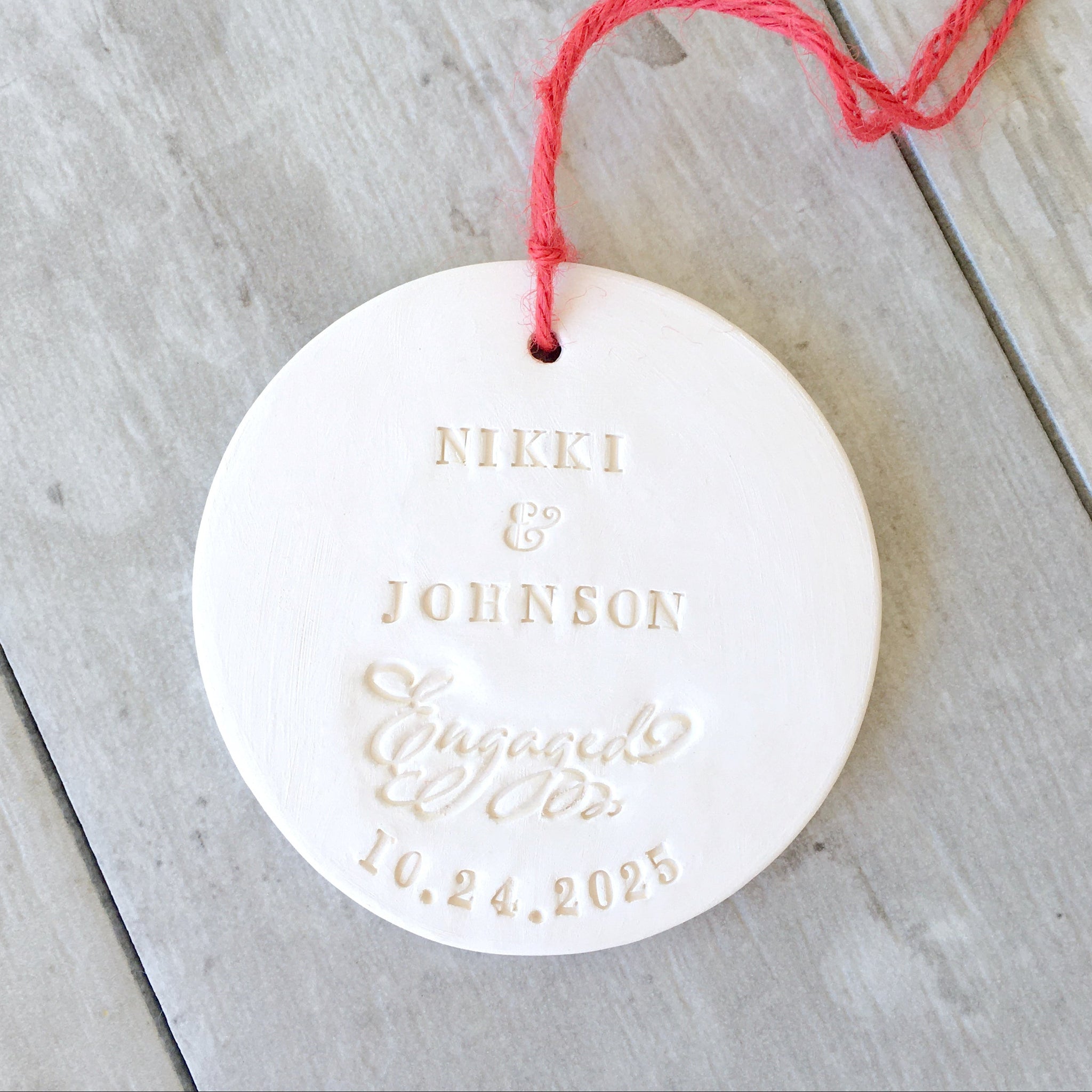 Handmade round white flat disc ceramic clay wedding ornament with a red hanging string, featuring embossed personalized typographic design reading 'NIKKI & JOHNSON', the word Engaged in script letters, and a date. Custom engagement gift for couple personalized with 2 first names and engaged date.