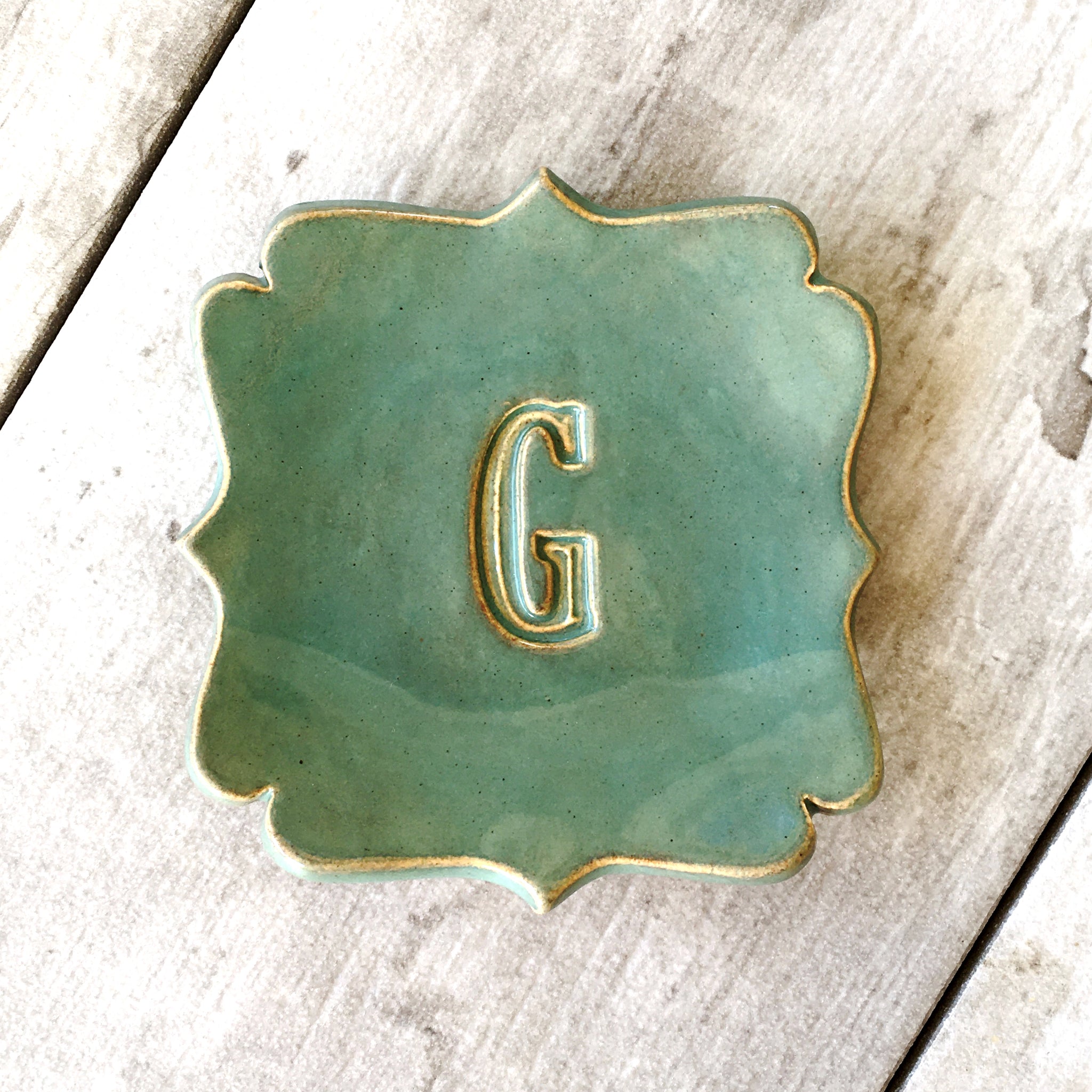 A scalloped ceramic monogram dish in a green glaze with rustic gold rim and a pressed letter 'G' in the center, photographed on a rustic whitewashed wood surface. Handmade ceramic curvy square tray personalized pottery spoon rest each artist created with your choice of hand engraved big name name letter & optional date, farmhouse kitchen utensil holder, curvy corner square ceramic tray gold tone letter & r