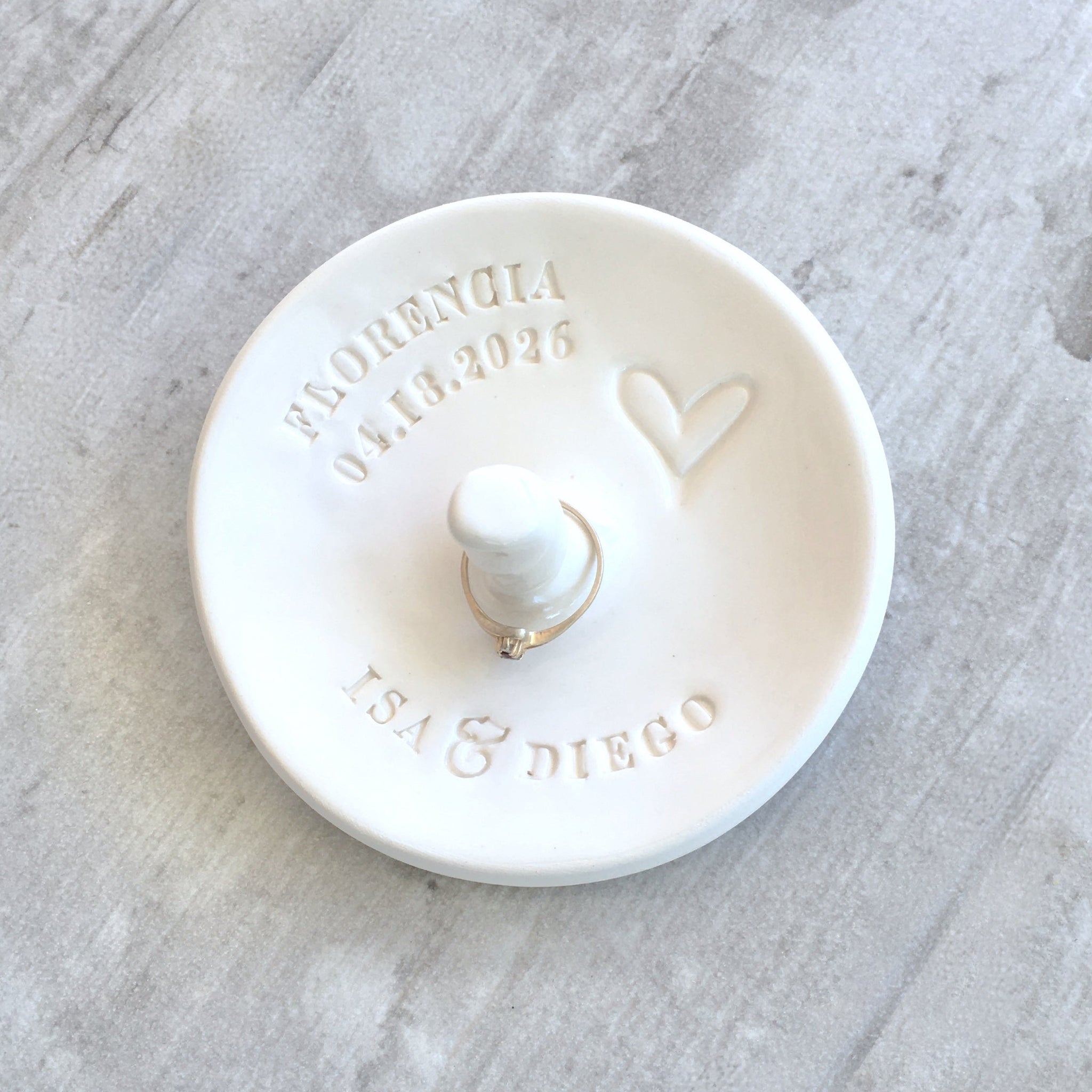 Artist handmade ceramic white gloss bowl with typographic design and centered jewelry post on wood surface. Custom pottery ring dish with cone includes hand engraved names and date, newlyweds ceremony plate, newly engaged couple gift proposal keepsake or party gift for bride to be, wedding memory wife anniversary gift for spouse. Tabletop or bedside table  jewelry storage for rings, bracelets, earrings, cuff links, pins.