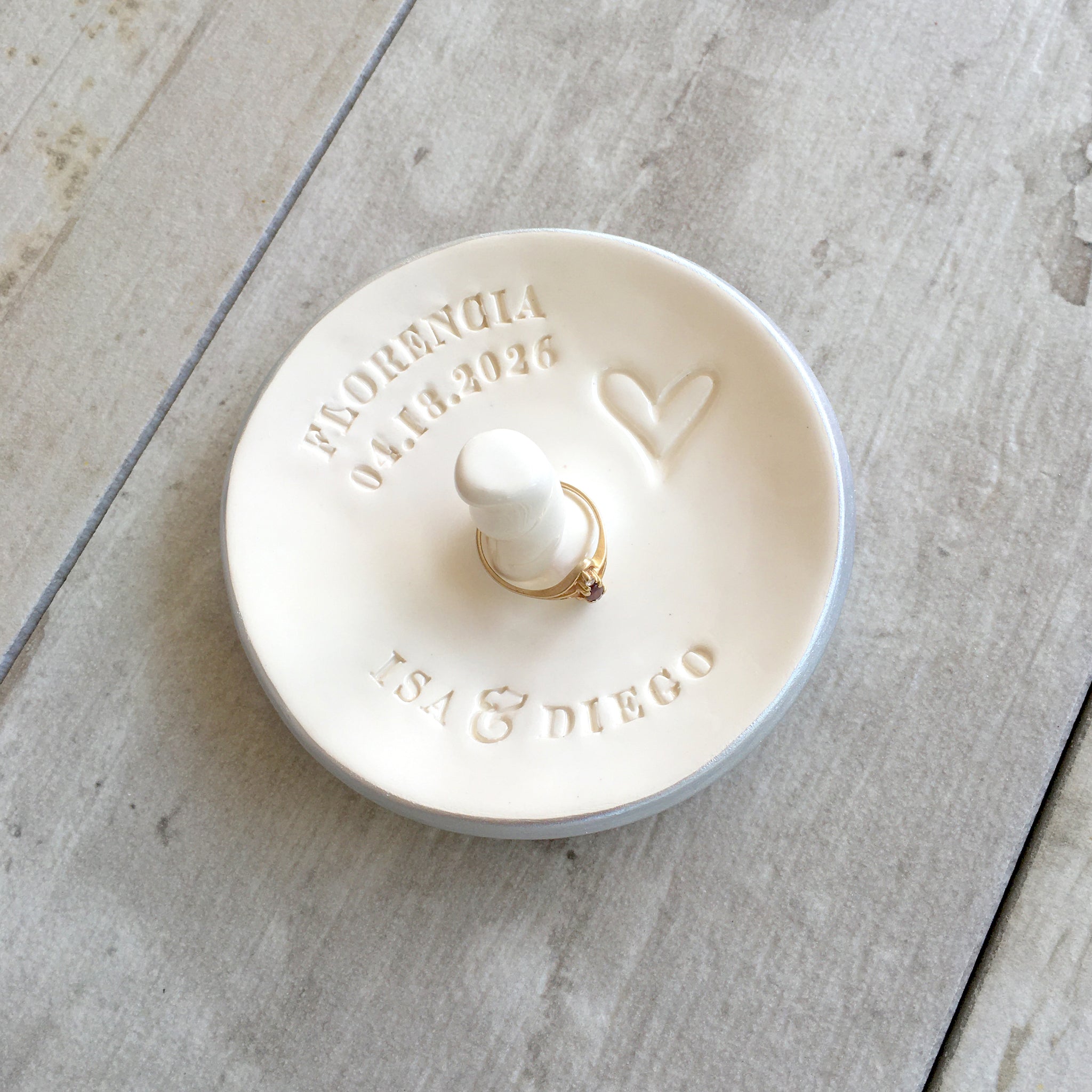 handmade ceramic white clay bowl pottery ring dish with central jewelry post, typographic design and silver rim on wood surface. Design includes hand engraved heart, 2 names, a location or city and a date. Custom wedding anniversary gift for couple. Ideal decorative tray, weddings memorial, engagement ring holder for proposal keepsake.