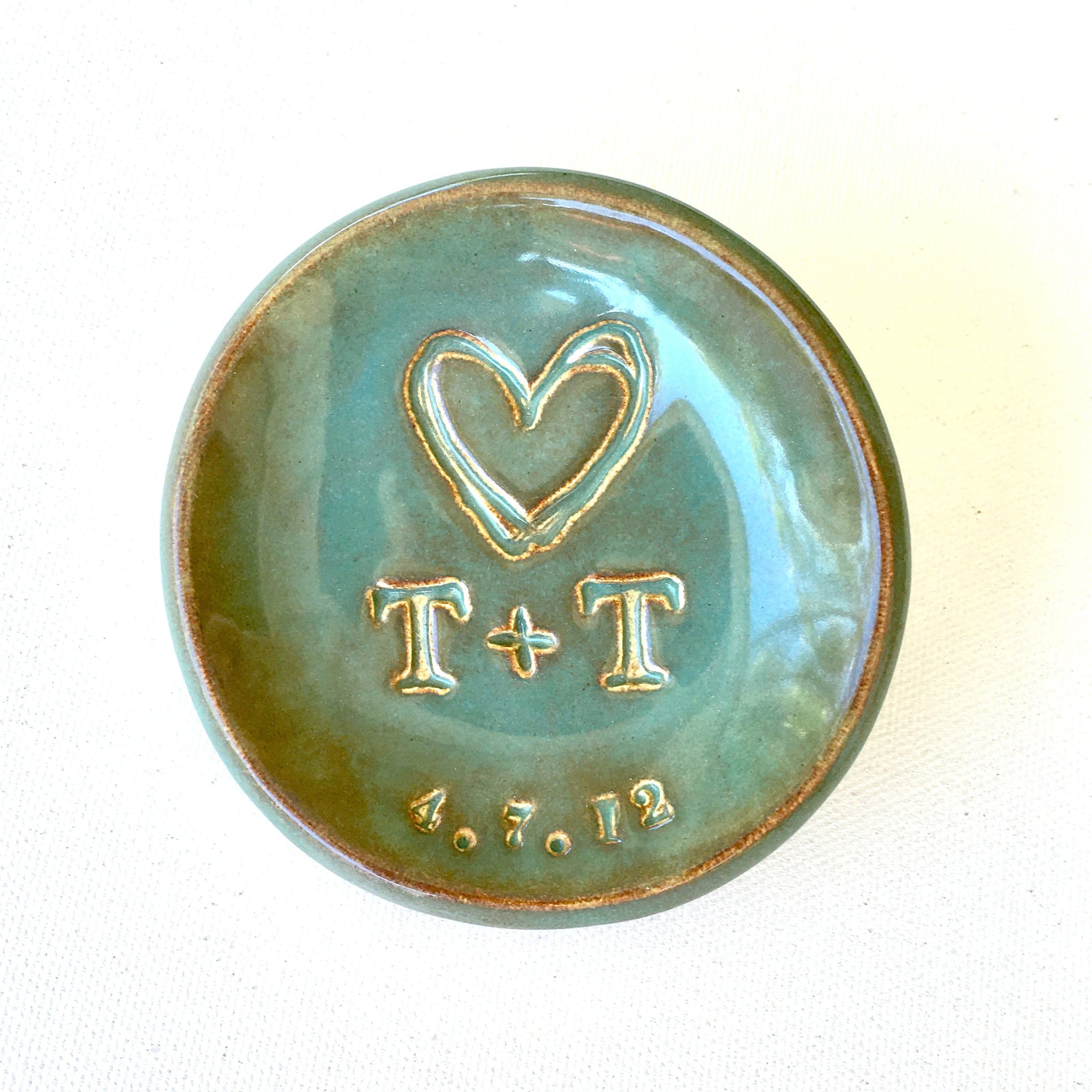 Photographed on white surface is a handmade ceramic personalized ring dish jewelry holder is rustic green decorative bowl with gold rim accents and typographic design pressed in clay.  Typographic design includes hand stamped 2 name initials, date and large heart. Name letters and date are accented in gold, customized wedding ring dish couple anniversary plate, jewelry holder