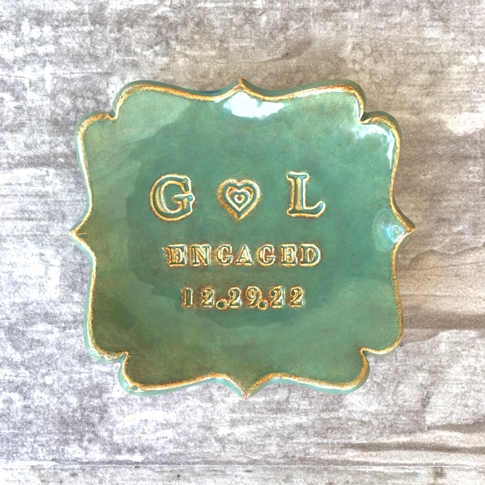handmade ceramic abstract square ring dish personalized jewelry holder in rustic green and gold glaze on wood surface. Typographic design includes a heart between 2 name initials, the word engaged & date. Modern farmhouse home decor tray. Unique engagement ring dish for engaged couple party gift and wedding rings tray for married anniversary keepsake. Ideal military wedding gift.