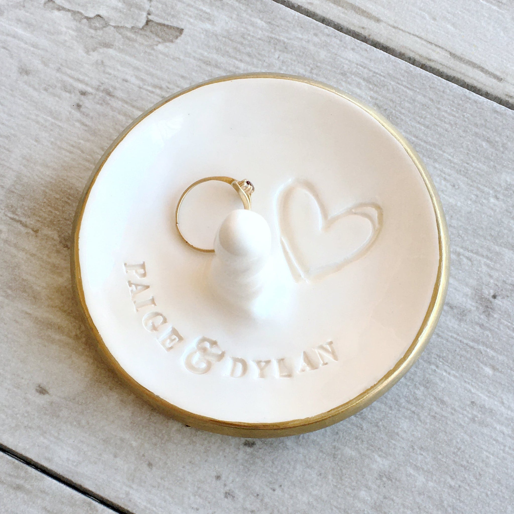 Handcrafted ceramic white bowl with gold rim, central rings peg, a ring & typographic design on gray wood surface. Personalized ring post dish with your 2 names & optional date, & outlined heart symbol pressed in clay. Ideal wedding ring dish engagement gift for couple, spouse anniversary pottery keepsake, partners dating girlfriend present. Jewelry storage for bracelets, earrings, necklaces on dresser or table top. Cute modern farmhouse home decor. Ideal engagement gift. 