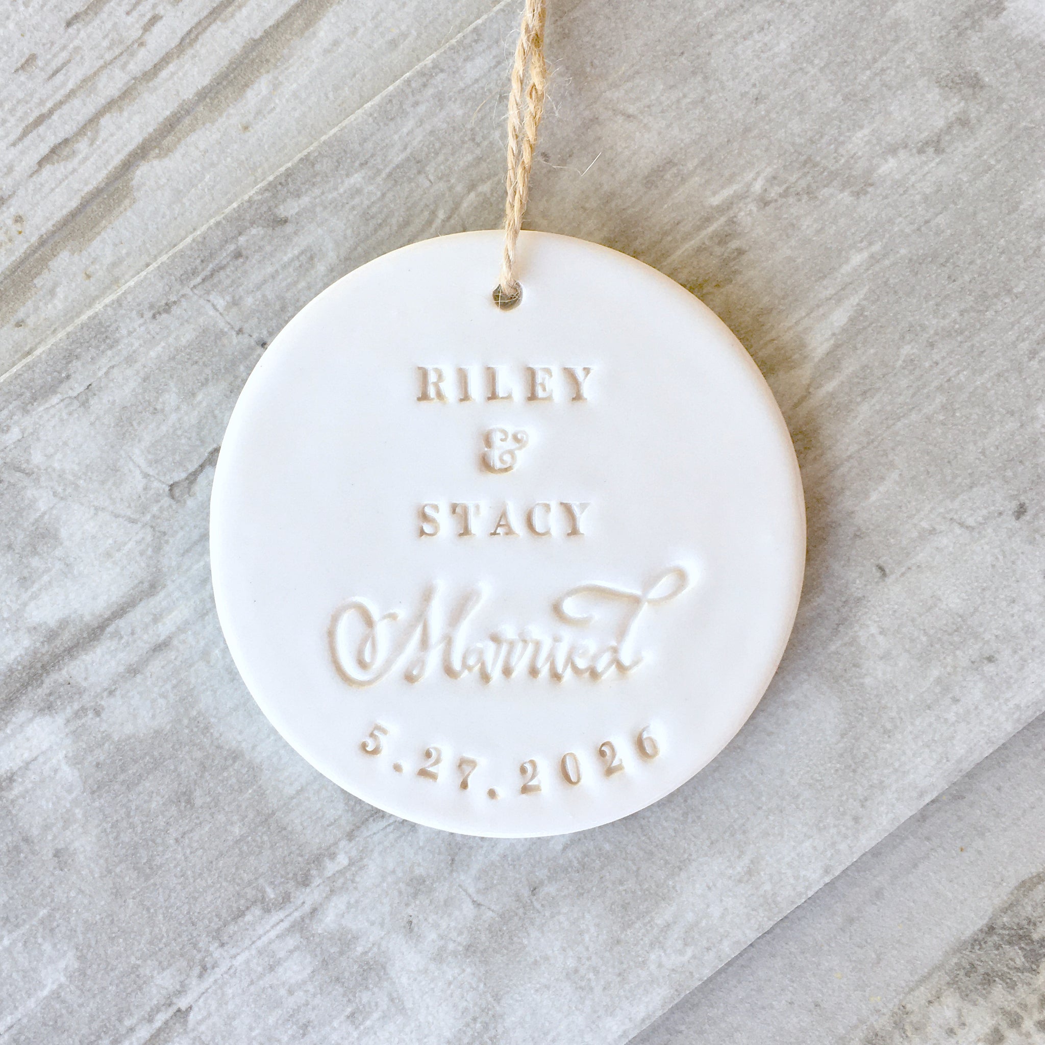 Handmade ceramic names ornament white round flat clay disc with smooth gloss surface, brown hang cord and typographic design in classic letter style.  Personalized weddings keepsake created with 2 first names, the word married in script, and a date. Custom pottery ornament hanging tree charm, weddings medallion for country farmhouse minimalist holiday decor marriage or couple proposal gift.