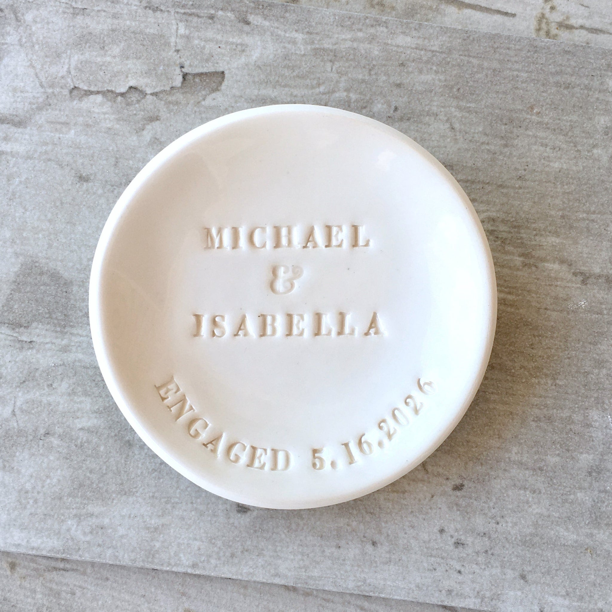 Handmade ceramic white clay bowl on wood surface with smooth gloss surface and typographic design in classic letter style. Personalized ring dish engagement gift with hand engraved 2 first names, the word engaged and a date, elegant white bowl with gold trim, unique bride to be gift.