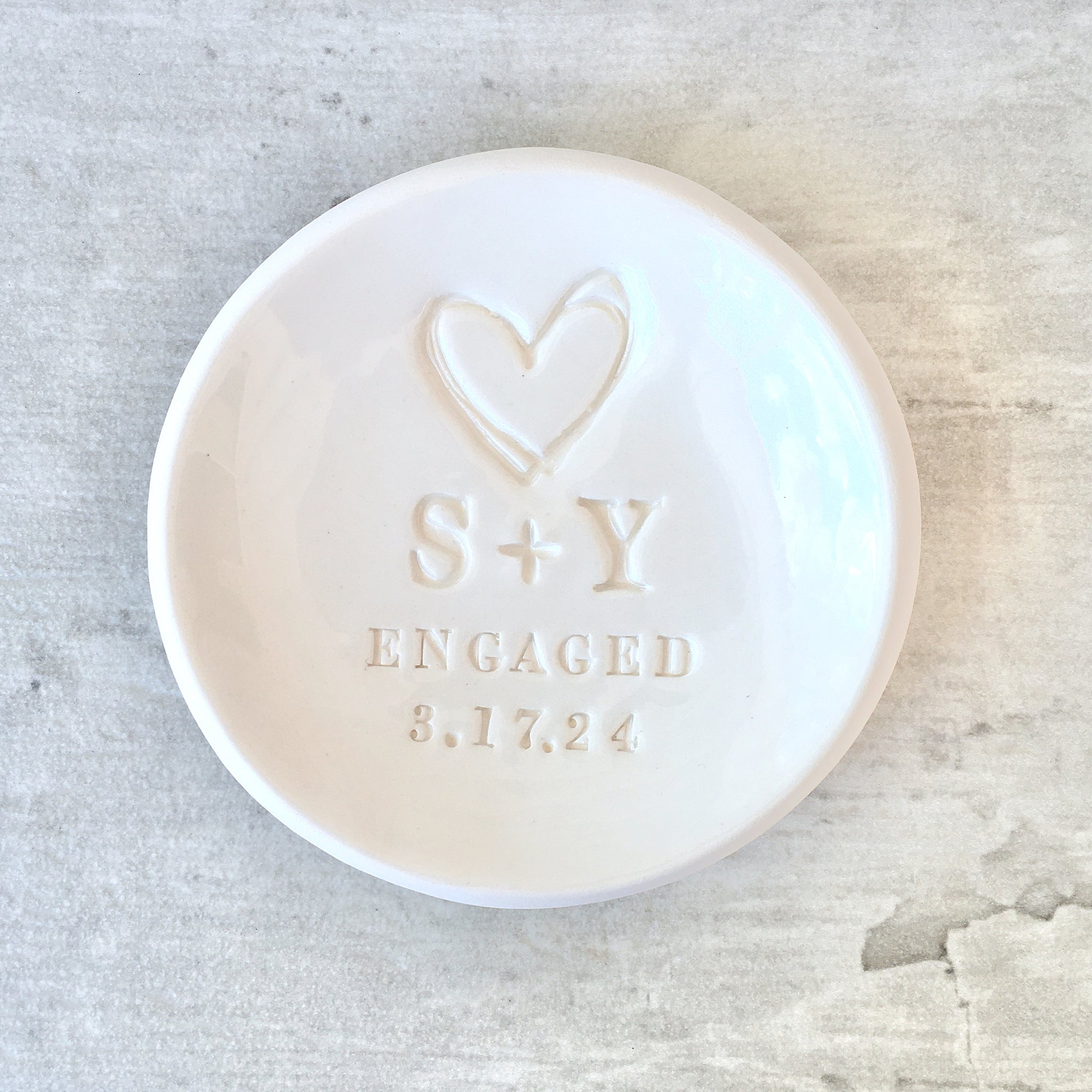 handcrafted pottery white gloss bowl with typographic design on wood surface. Artist created personalized ring dish engagement ring holder engaged couple gift with large heart, 2 name initials, the word engaged and a date pressed in clay. Modern minimalist home decor tray jewelry storage. 