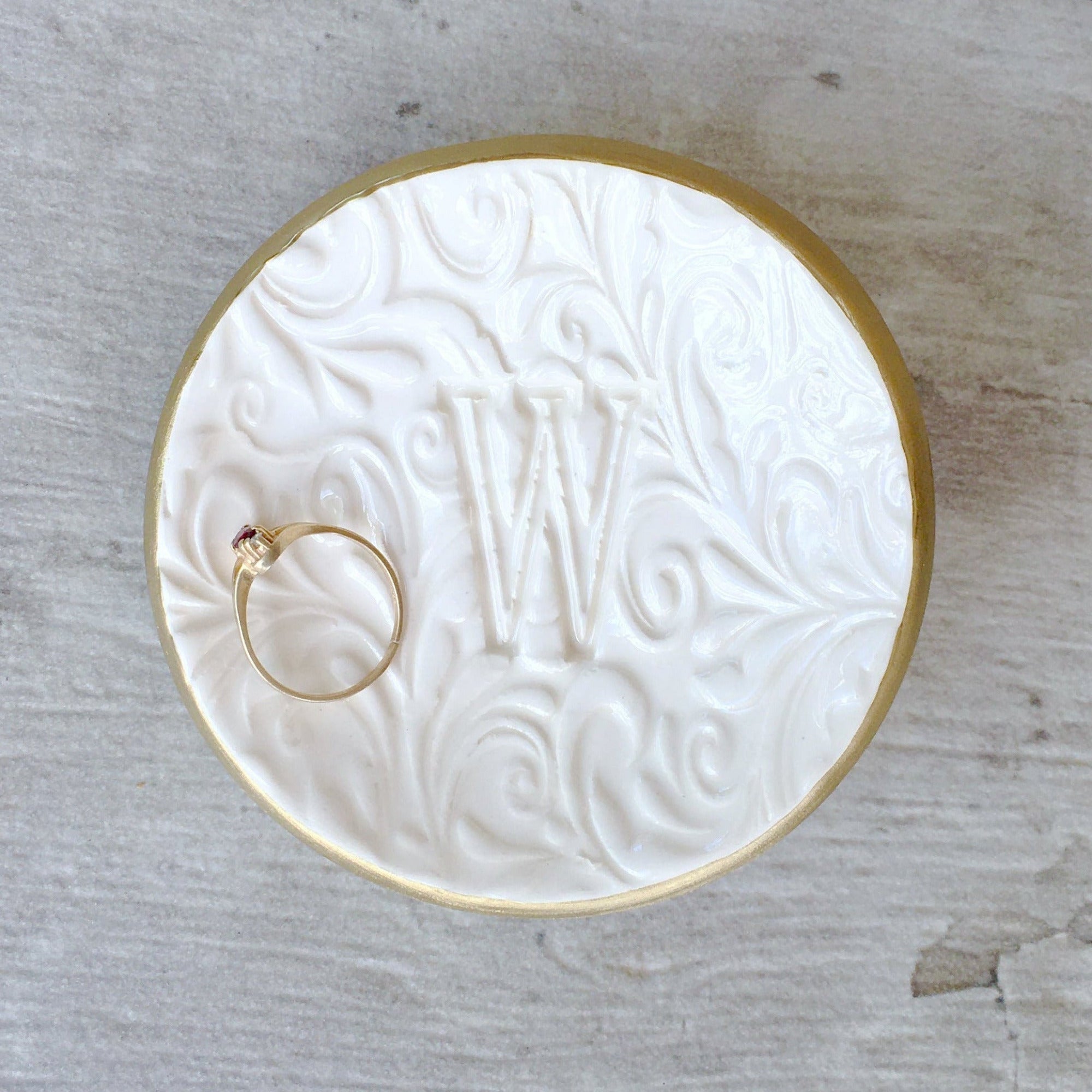 Handmade ceramic white gloss bowl with gold rim & hand pressed curvy floral decorative pattern surrounding a large initial letter on a wooden surface, dish is holding a ring. Unique personalized ring holder jewelry tray with a name letter pressed in clay. Ideal wife gift for women, daughters, bridesmaid thank you, medical staff present, wedding ring holder, engagement ring bowl, teen, girlfriend women's birthday. Storage for rings, bracelets, earrings, necklaces by sink, dresser, tabletop decor.