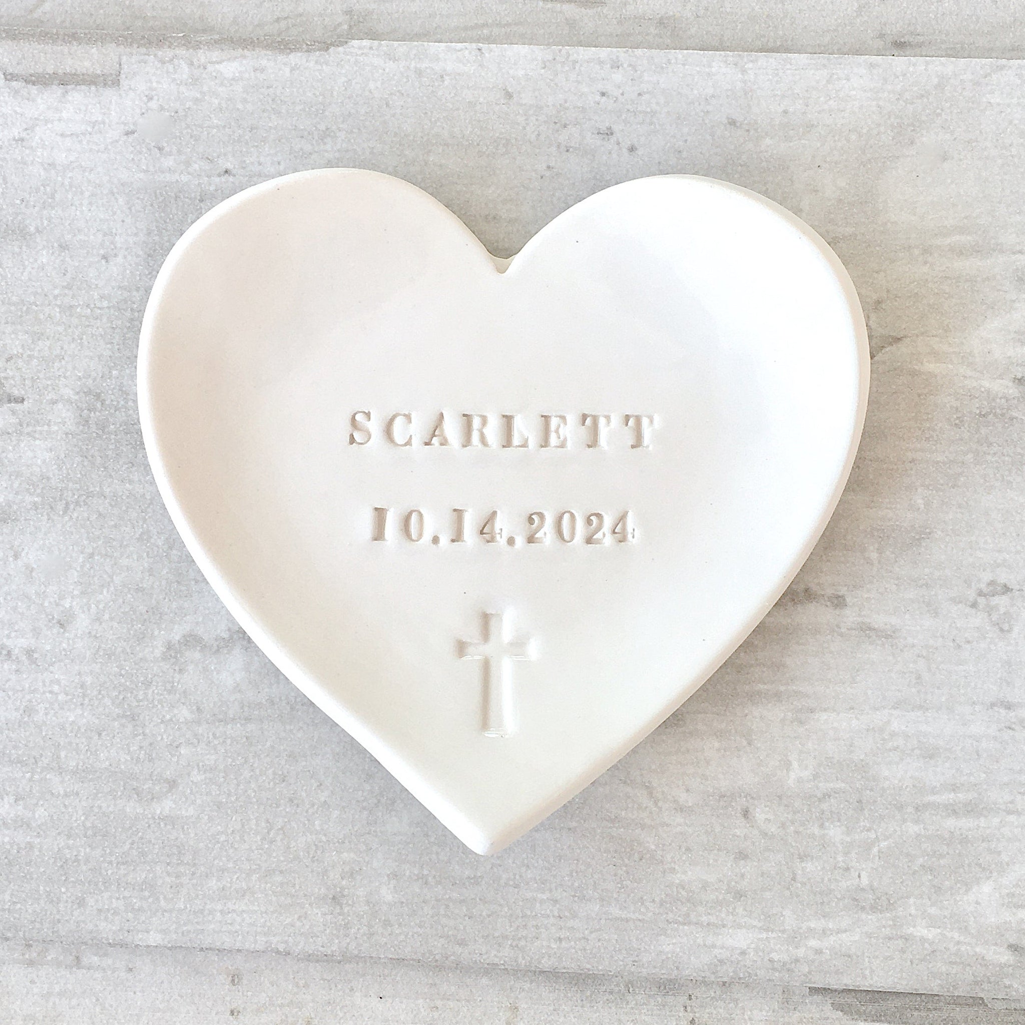 Handmade pottery, a heart-shaped white ceramic dish with cross and personalized embossed text includes your name and a date. Photographed on wood surface. Ideal for rosary holder or sacraments gifts: first communion baptism confirmation