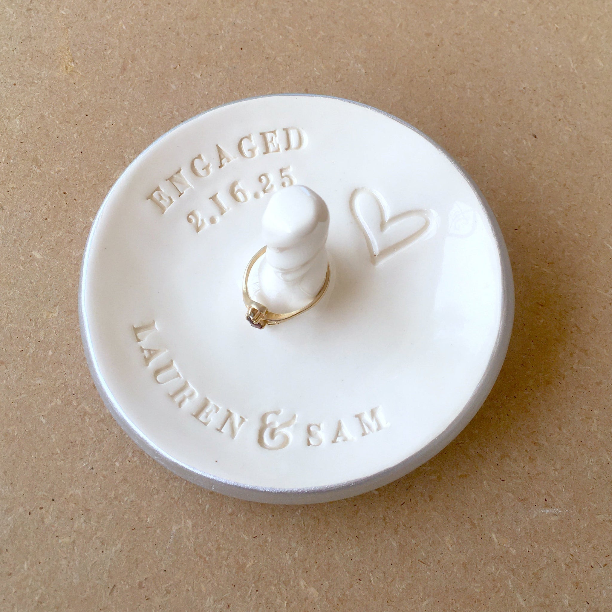 Handmade ceramic gloss white bowl with classic silver trim shown on wood surface, custom engagement gift personalized ring dish with post, includes engraved heart, names and optional word engaged and date, also feature a central jewelry display cone to safely secure bracelets, rings, necklaces. Newly engaged couple gift, bridal shower present, future mr and mrs wedding keepsake.