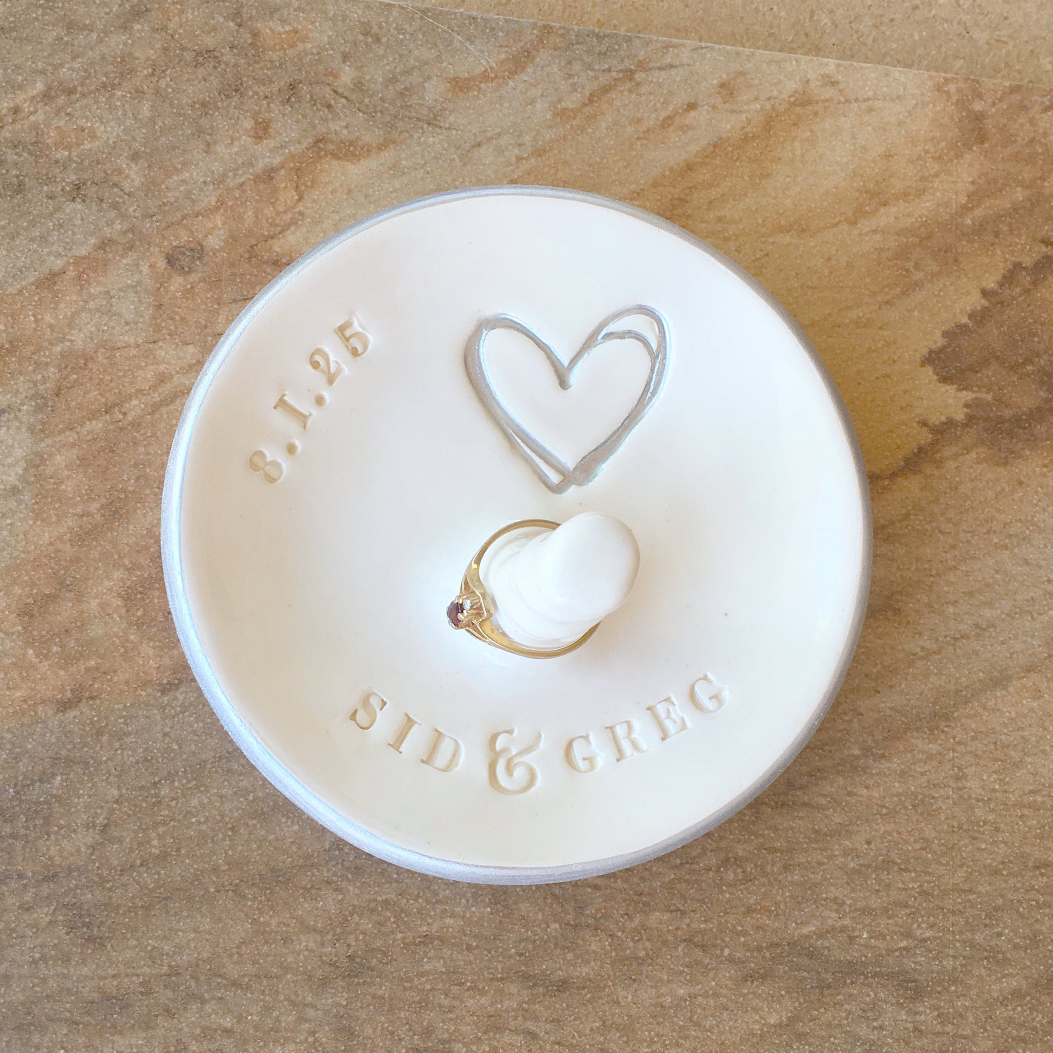 Handmade ceramic modern silver rim white bowl personalized ring dish with jewelry post and typographic design on wood surface. Made to order couple jewelry display engagement ring holder features hand engraved names date and large heart, heart is painted silver. Engaged bride to be proposal gift.
