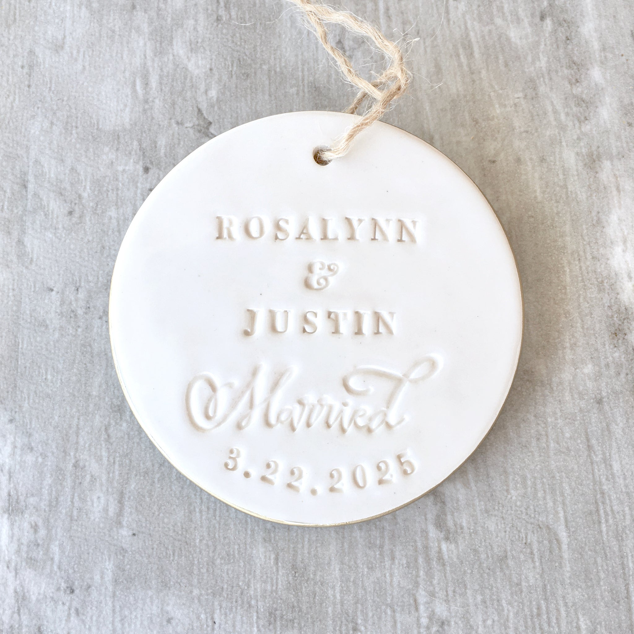 Handmade ceramic names ornament white round flat clay disc with smooth gloss surface, brown hang cord and typographic design in classic letter style.  Personalized weddings keepsake created with 2 first names, the word married in script, and a date. Custom pottery ornament hanging tree charm, weddings medallion for country farmhouse minimalist holiday decor marriage or couple proposal gift.