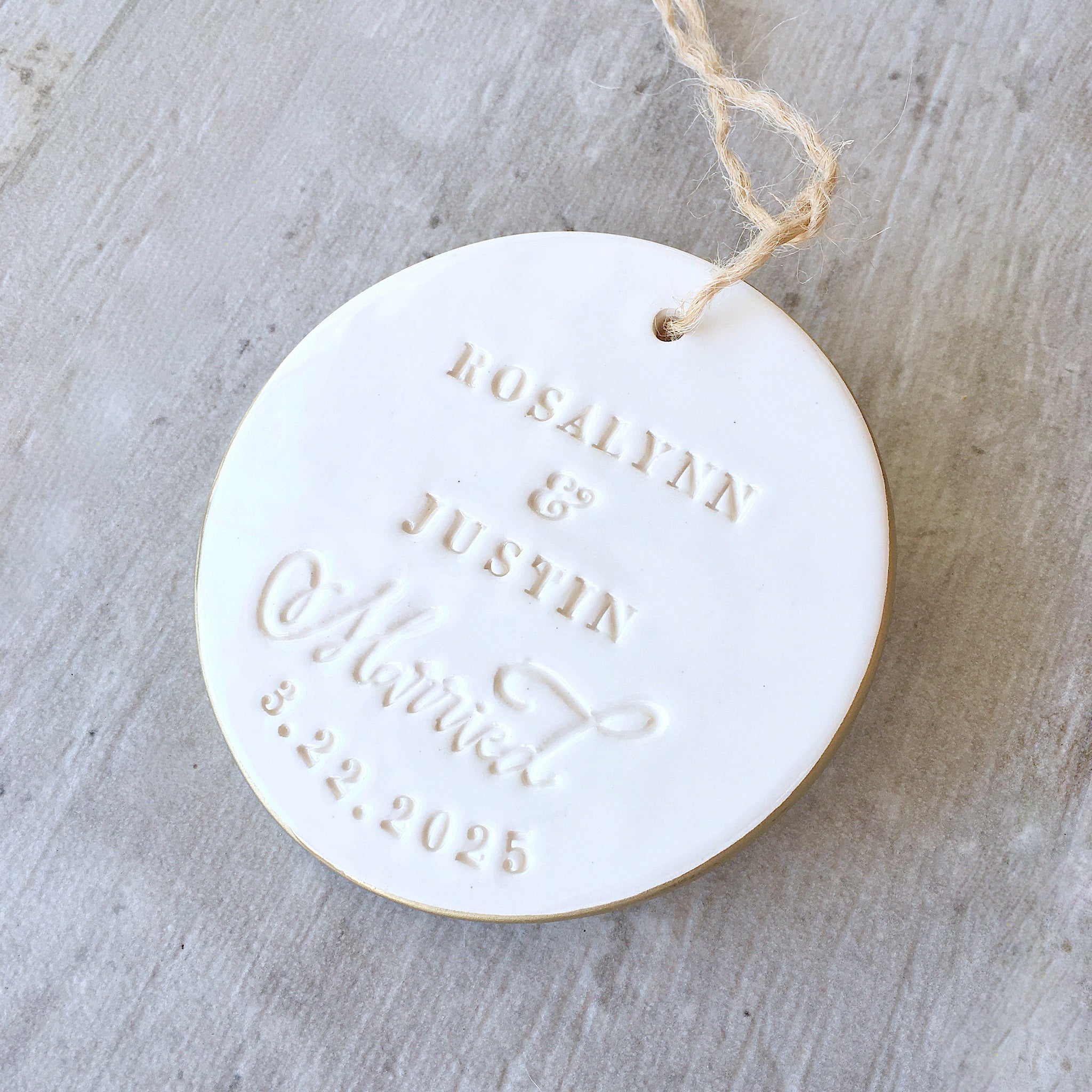 Handmade ceramic names ornament white round flat clay disc with gold rim & smooth gloss surface, brown hang cord and typographic design in classic letter style.  Personalized weddings keepsake created with 2 first names, the word married in script, and a date. Custom pottery ornament hanging tree charm, weddings medallion for country farmhouse minimalist holiday decor marriage or couple proposal gift.