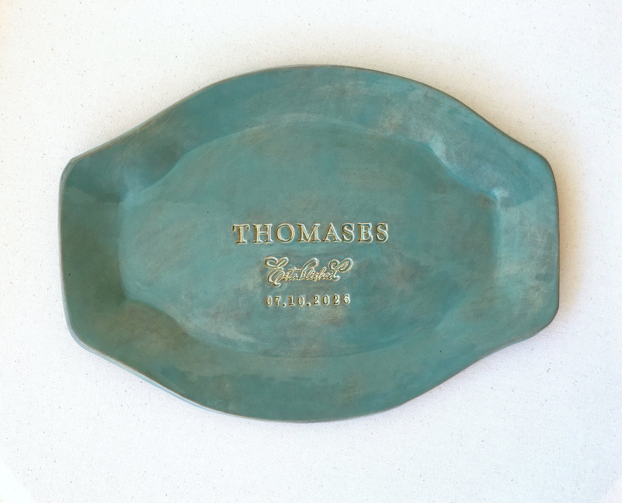 handmade ceramic platter oval tray with rustic green glaze finish that has gold and brown marbling, dish on a light surface. Keepsake plate has with hand engraved  family name, the word established and a date, golden accents on trim and gold lettering. Personalized pottery tray with handles, unique wedding anniversary gift for couple, ceremony plate.