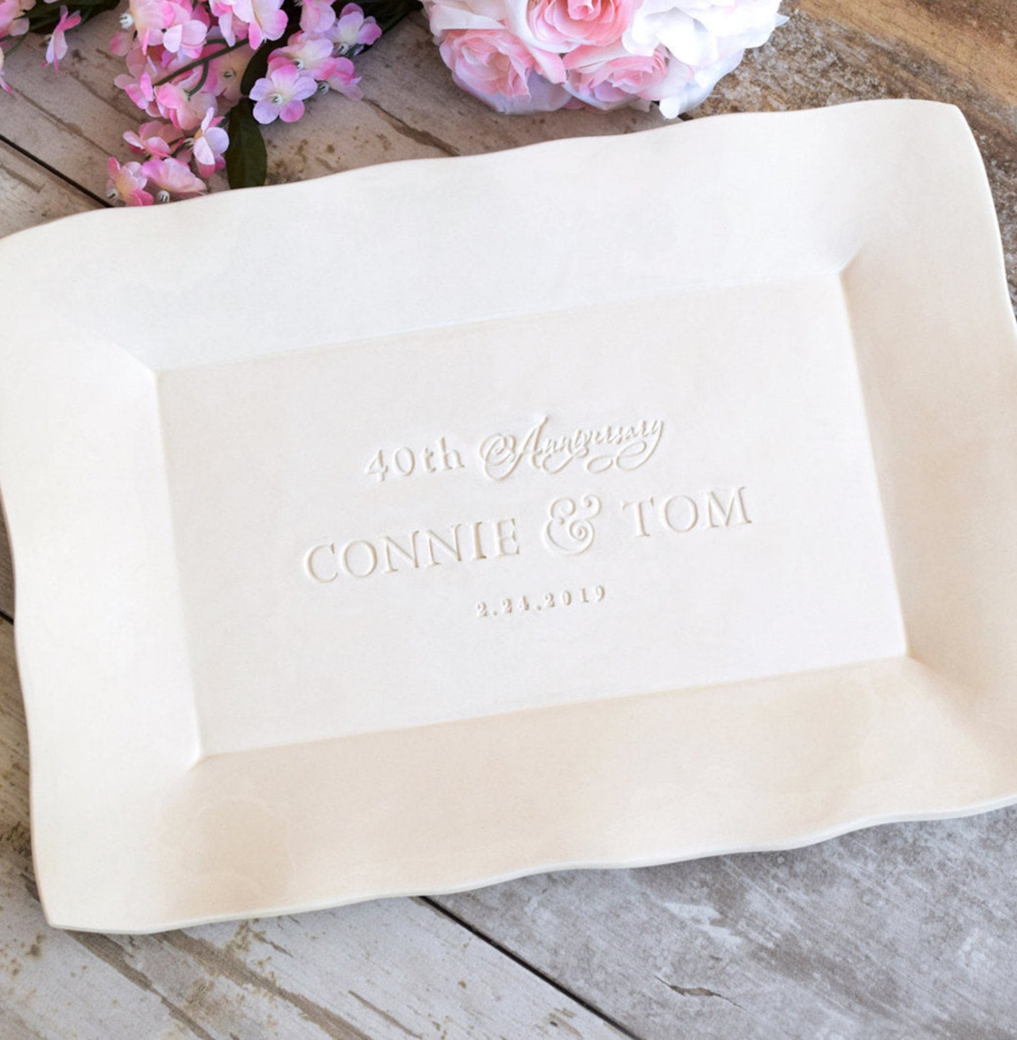 handmade ceramic anniversary platter with golden gloss finish, typographic design hand stamped in clay. Large rectangular wedding tray with wavy sides on wood surface with pink flowers. Design features hand pressed letters and numbers: anniversary year, the word anniversary in calligraphy script, 2 first names and a date. Custom gift for couple, parents family celebration. Ideal spouse milestone keepsake, 8 year or 9th anniversary pottery, grandparents 50th