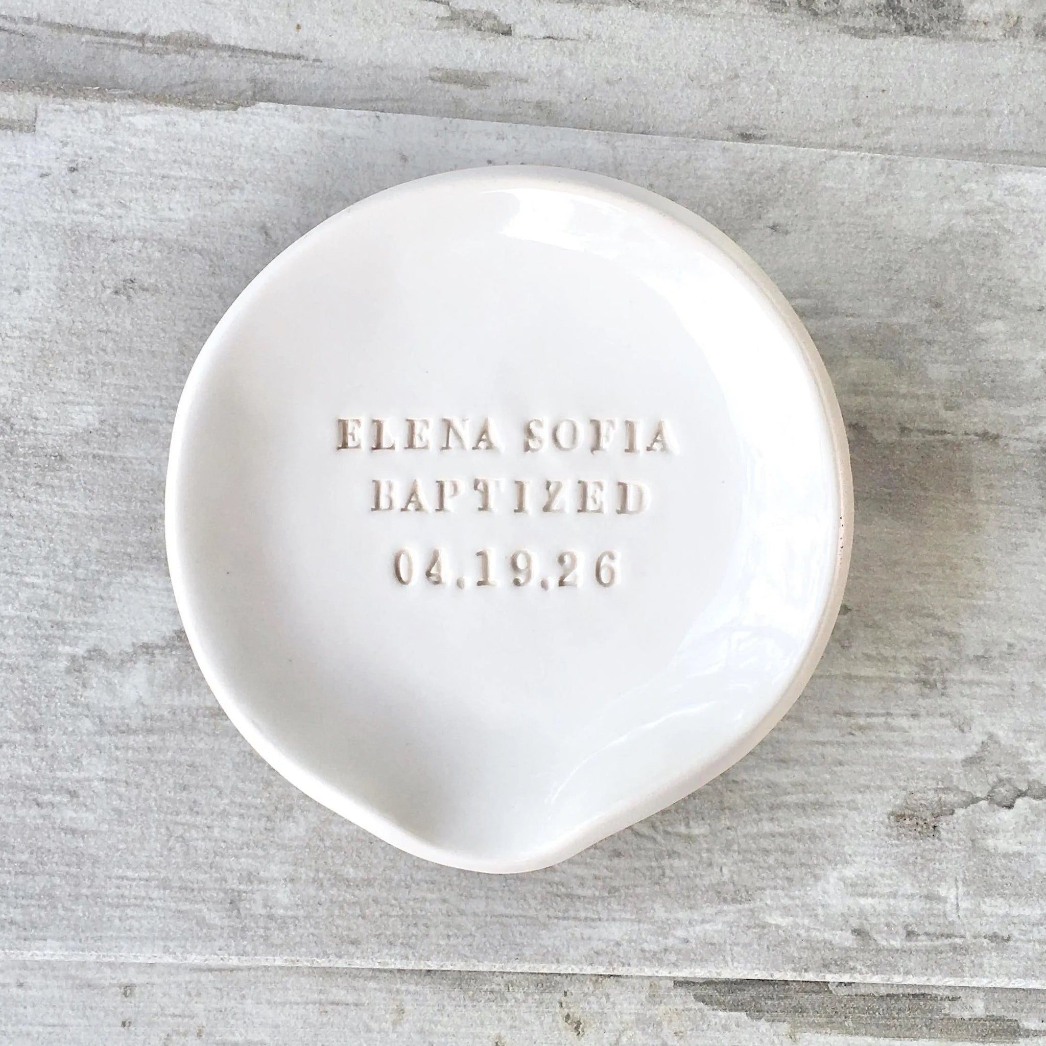 A white handmade ceramic personalized baptismal font water bowl for christening with hand pressed lettering: a first and middle name, the word 'baptized' and a date. Photographed on wood surface.