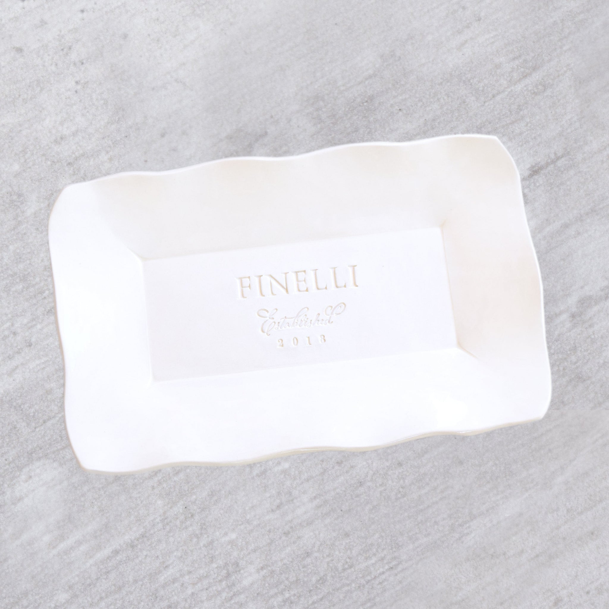 handmade ceramic white platter with wavy sides on wood surface. Large rectangular platter has gloss finish, typographic design hand stamped in clay: family name, date & the word established in script font. Country chic home kitchen serving tray. Handcrafted on wedding day memory keepsake souvenir, spouse anniversary tray, serving tray. Unique weddings gift for family, parents, coworker, employee retirement & event memento. White on white minimalist decor hand engraved clay.