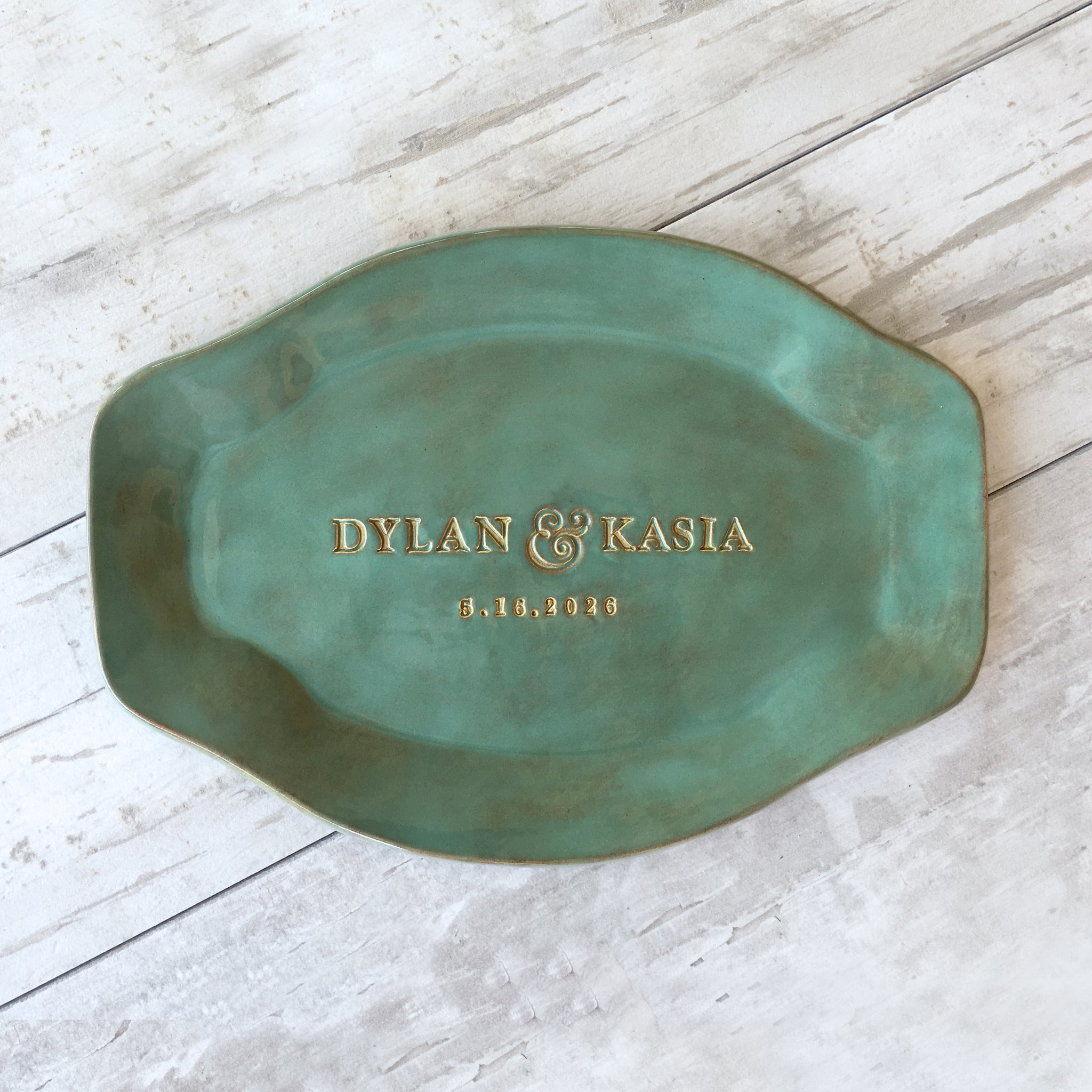 Green handmade ceramic personalized platter with typographic design. Sample dish shown includes names 'Dylan & Kasia' and a date on a light wooden surface. Oval serving tray with rustic green glaze finish that has gold and brown marbling. Keepsake plate created with hand pressed names and date, golden accents on trim and lettering. Unique event plate, retirement memento, wedding anniversary gift for couple, ceremony plate. 