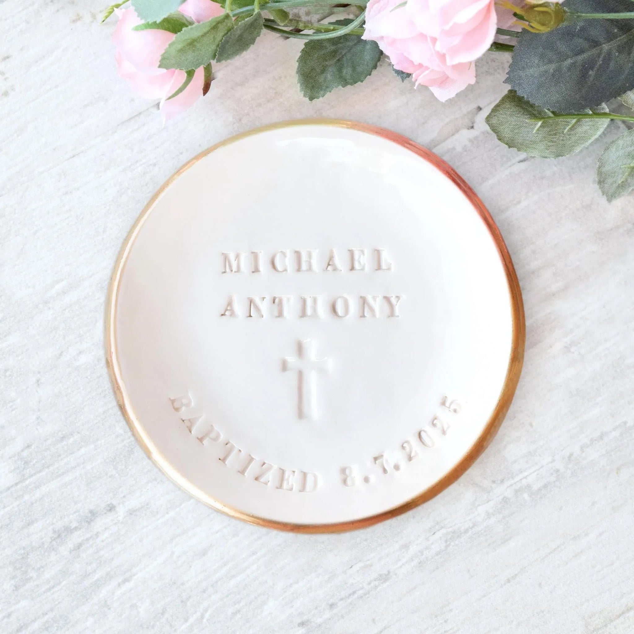 Handmade ceramic white bowl with smooth surface & gold rim has hand stamped typographic design and Christian cross symbol on wood surface with pink flowers. Water baptism keepsake heirloom Catholic sacraments ceremony gift. Includes first and middle name, the word baptized, a date & a cross. Ideal water baptismal christening bowl. Spiritual gift for new parents or Christ follower, Catholic child sacrament keepsake. Unique trinket tray, rosary tray, jewelry display, table top decor, small candle holder.