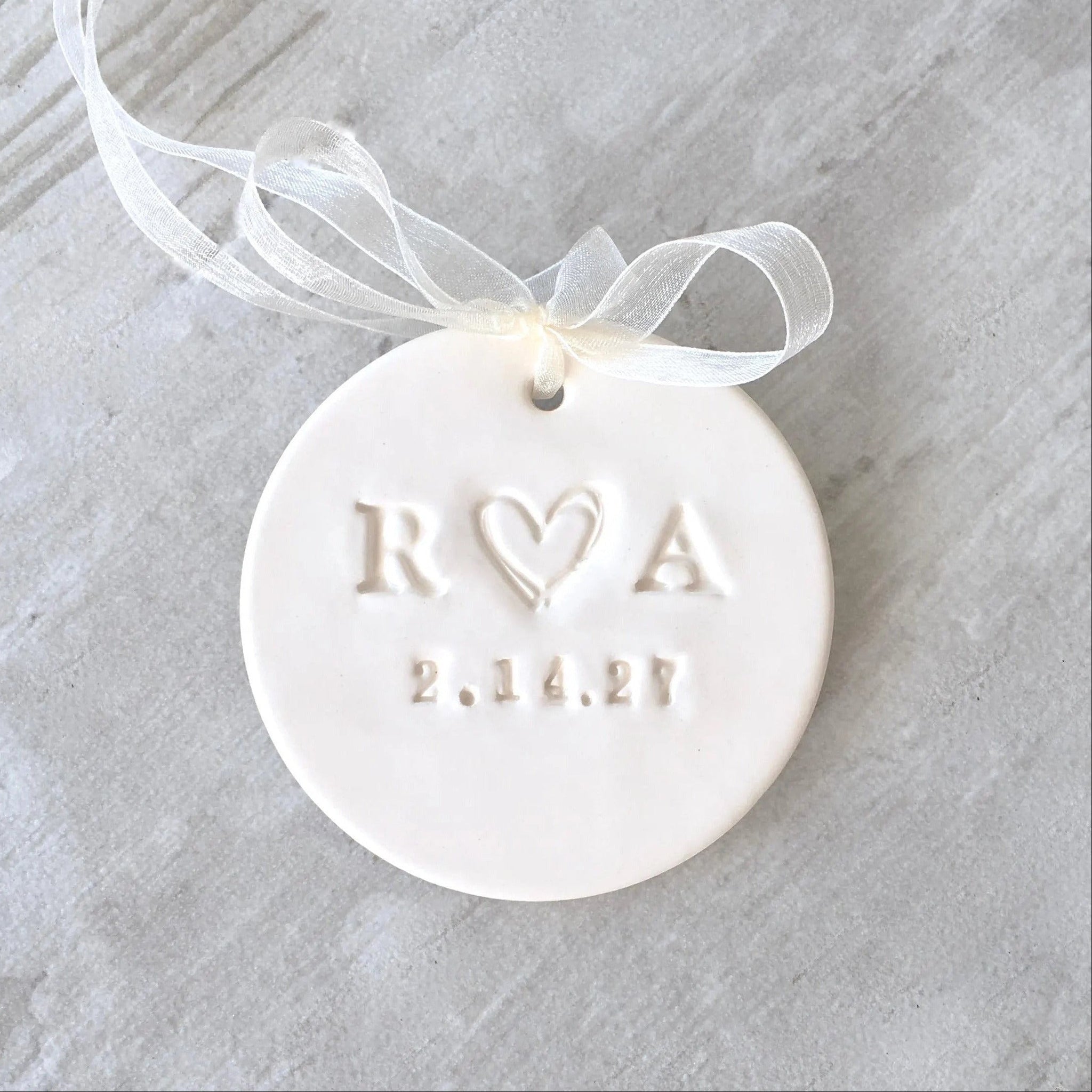 handmade ceramic ornament is white flat clay disc with smooth surface and typographic design of 2 name initials and a date. Hanging ornament has a sheer ribbon tied into a bow for hanging & is displayed on gray wood. Personalized marriage ornament or proposal bridal ornament for engaged couple first Christmas keepsake. Christmas tree decor holiday decoration. Modern farmhouse style charm, unique holiday stocking stuffer, ideal for newly engaged couple party or married newlyweds gift.