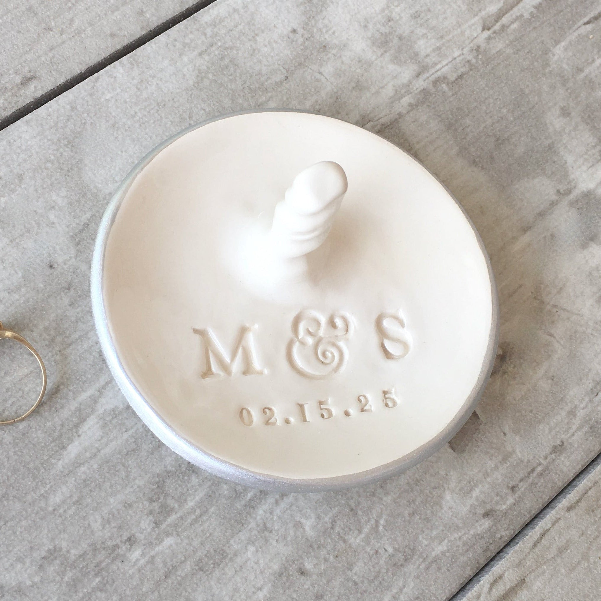 Handmade ceramic round white dish with centered rings post & silver rim edge on a wood surface with ring. Custom engagement gift personalized wedding ring dish with post, includes hand engraved name initials & a date, also features a central jewelry display cone to safely secure bracelets, rings, necklaces. Newly engaged couple gift, bridal shower present, future mr and mrs ceremony or anniversary keepsake.