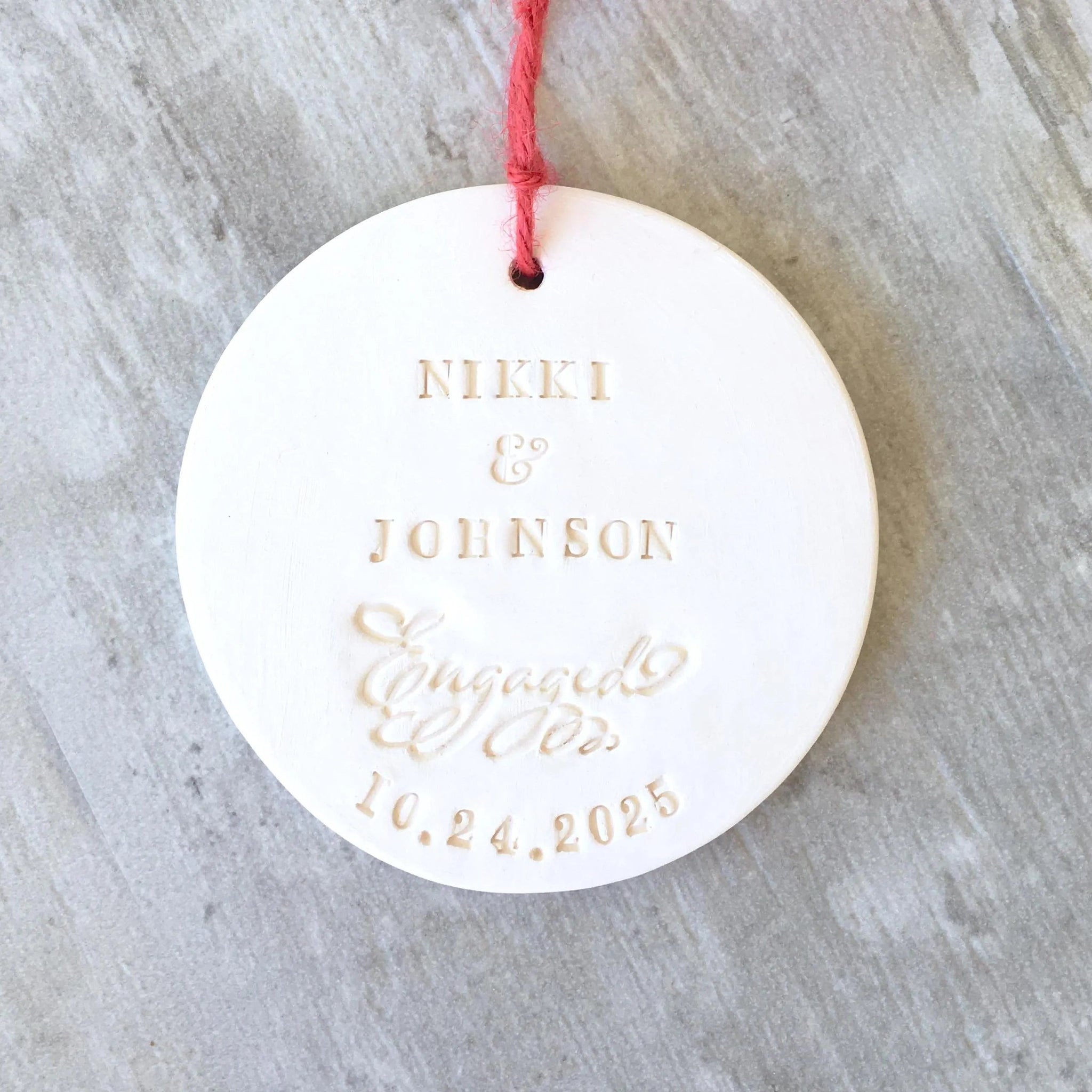Handmade ornament ceramic clay charm white round flat disc with smooth surface, red hang cord and typographic design in classic letter style.  Personalized weddings engagement keepsake created with 2 first names, the word engaged in script, and a date. Custom pottery ornament hanging tree medallion, weddings keepsake for country farmhouse minimalist holiday decor marriage or couple proposal gift.