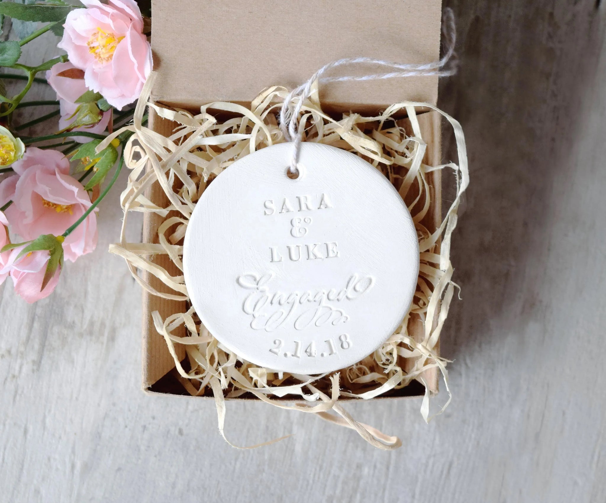 Handmade ornament ceramic clay charm white round flat disc with smooth surface, tan hang cord and typographic design in classic letter style in brown gift box, gray surface with pink flowers.  Personalized weddings engagement keepsake created with 2 first names, the word engaged in script, and a date. Custom pottery ornament hanging tree medallion, weddings keepsake for country farmhouse minimalist holiday decor marriage or couple proposal gift.