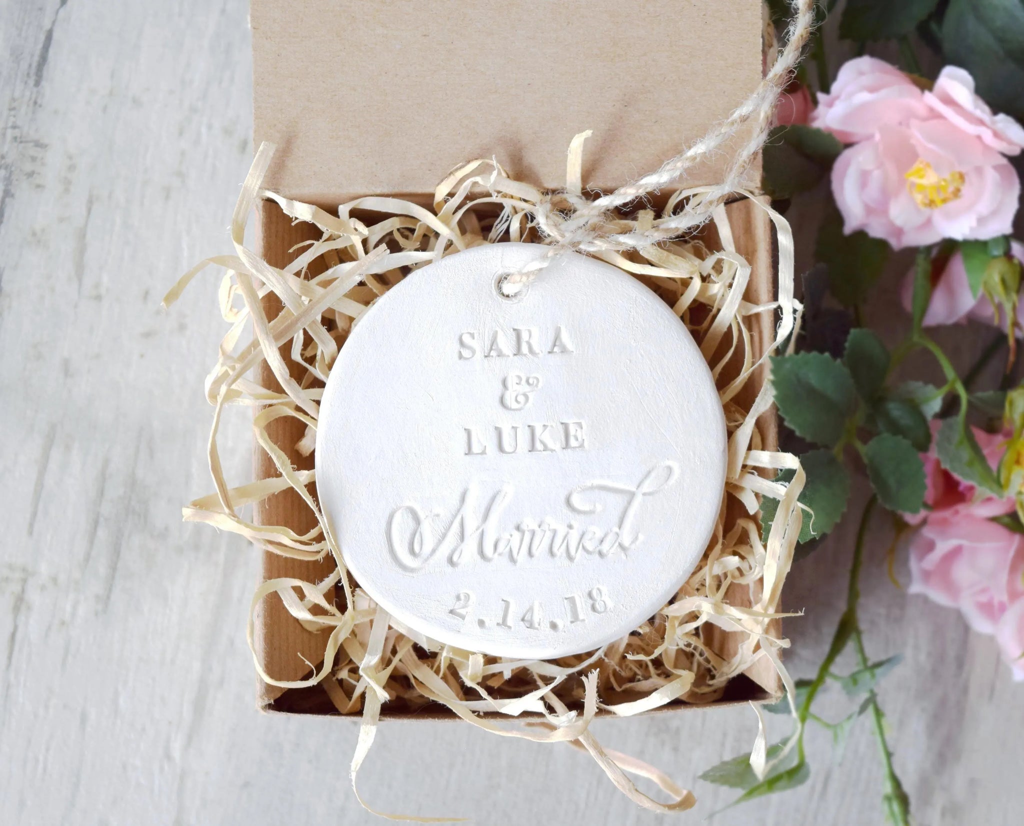Handmade ornament ceramic clay charm white round flat disc with smooth surface, tan hang cord and typographic design in classic letter style in gift box on gray surface with pink flowers.  Personalized weddings engagement keepsake created with 2 first names, the word married in script, and a date. Custom pottery ornament hanging tree medallion, weddings keepsake for country farmhouse minimalist holiday decor marriage or couple proposal gift.