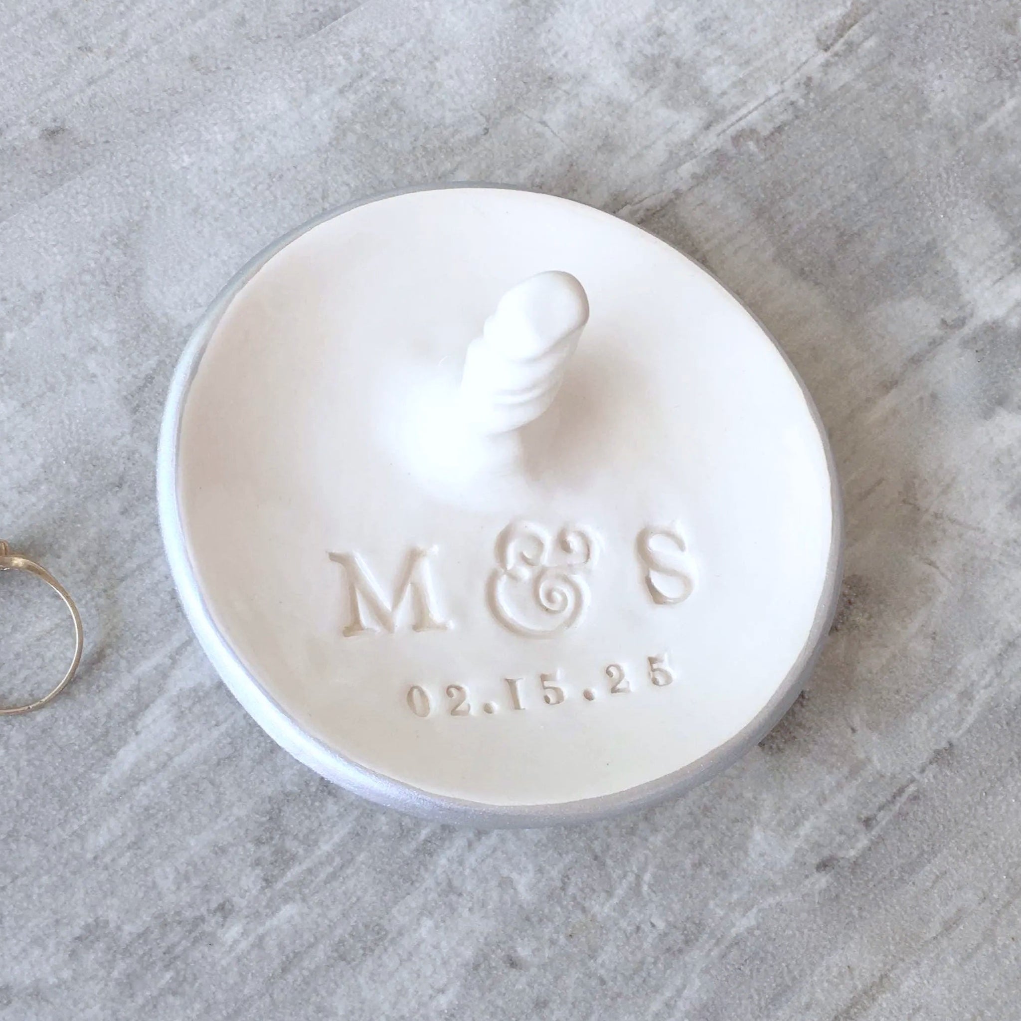 A round white handcrafted ceramic ring holder with central stand, embossed with the 2 initials and date. Handmade ceramic round white dish with centered rings post & silver rim edge on a wood surface with ring. Custom engagement gift personalized wedding ring dish with post, includes hand engraved name initials & a date, also features a central jewelry display cone to safely secure bracelets, rings, necklaces. Newly engaged couple gift, bridal shower present, future mr mrs ceremony or anniversary keepsake.