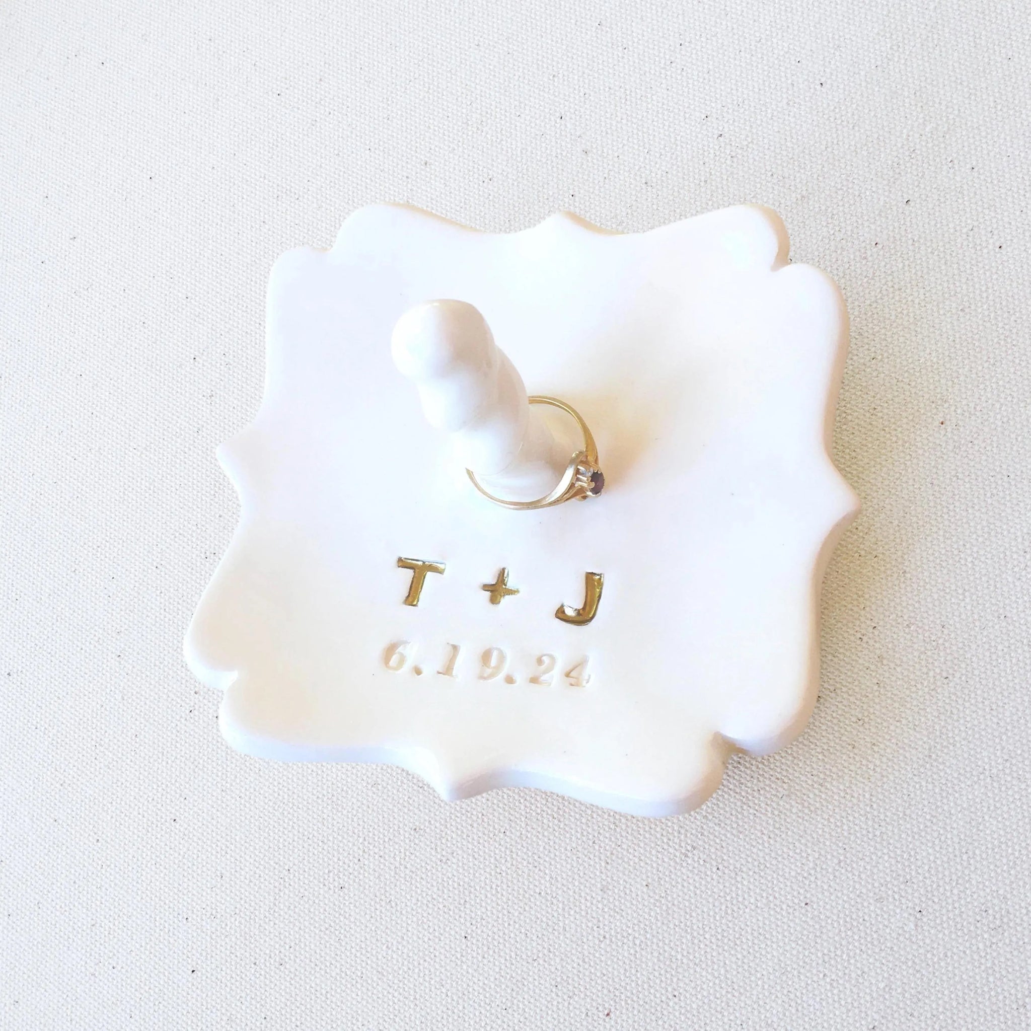 Handmade ceramic square tray with curvy sides, Typographic design and jewelry post  on light textured surface. Personalized ring holder created with your engraved 2 name initials, a plus sign and a date. Initials are painted gold. Ideal custom engagement ring holder wedding keepsake with central jewelry post. Customized ceremony or celebration display home decor for couple. Useful to secure rings bracelets, necklaces. Daughter bridal memento, or spouse anniversary gift.